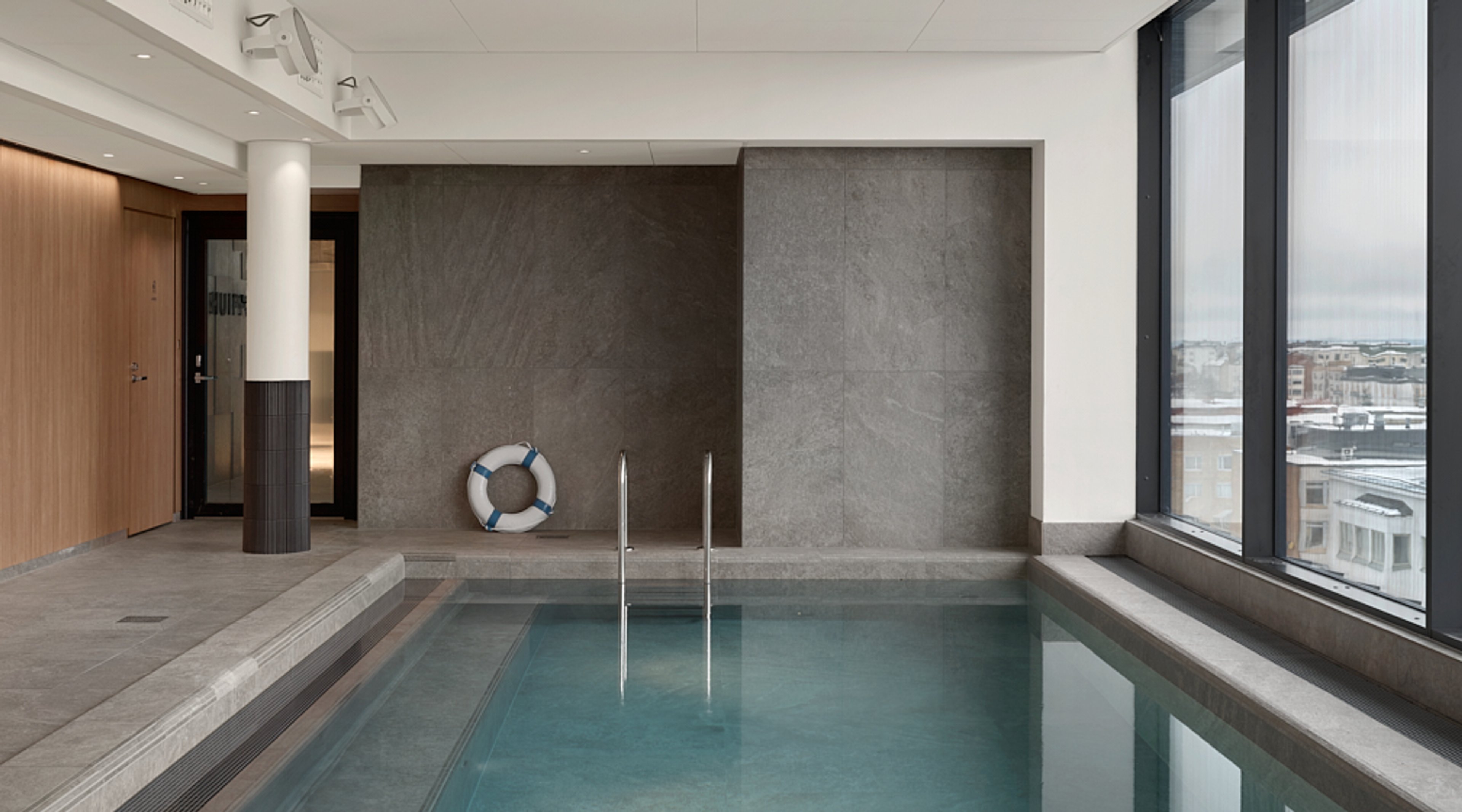 Moderni sisäuima-allas suurilla ikkunoilla, joista avautuu kaupunkimaisema. Altaassa metallinen käsikaide ja reunalla pelastusrengas.