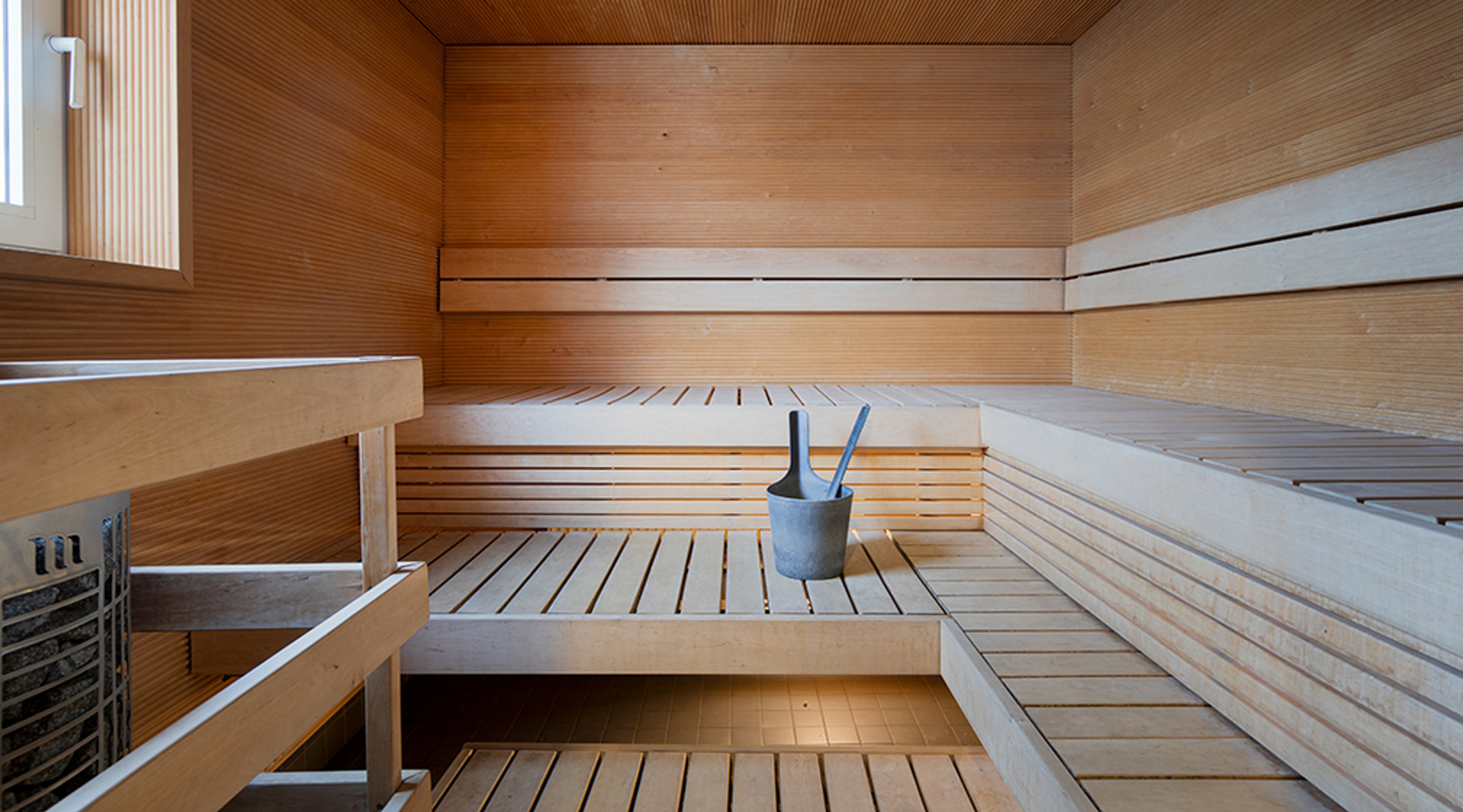 Sisäkuva saunasta puisilla penkeillä ja seinäpaneeleilla. Keskellä kiulu ja saunakauhoja.