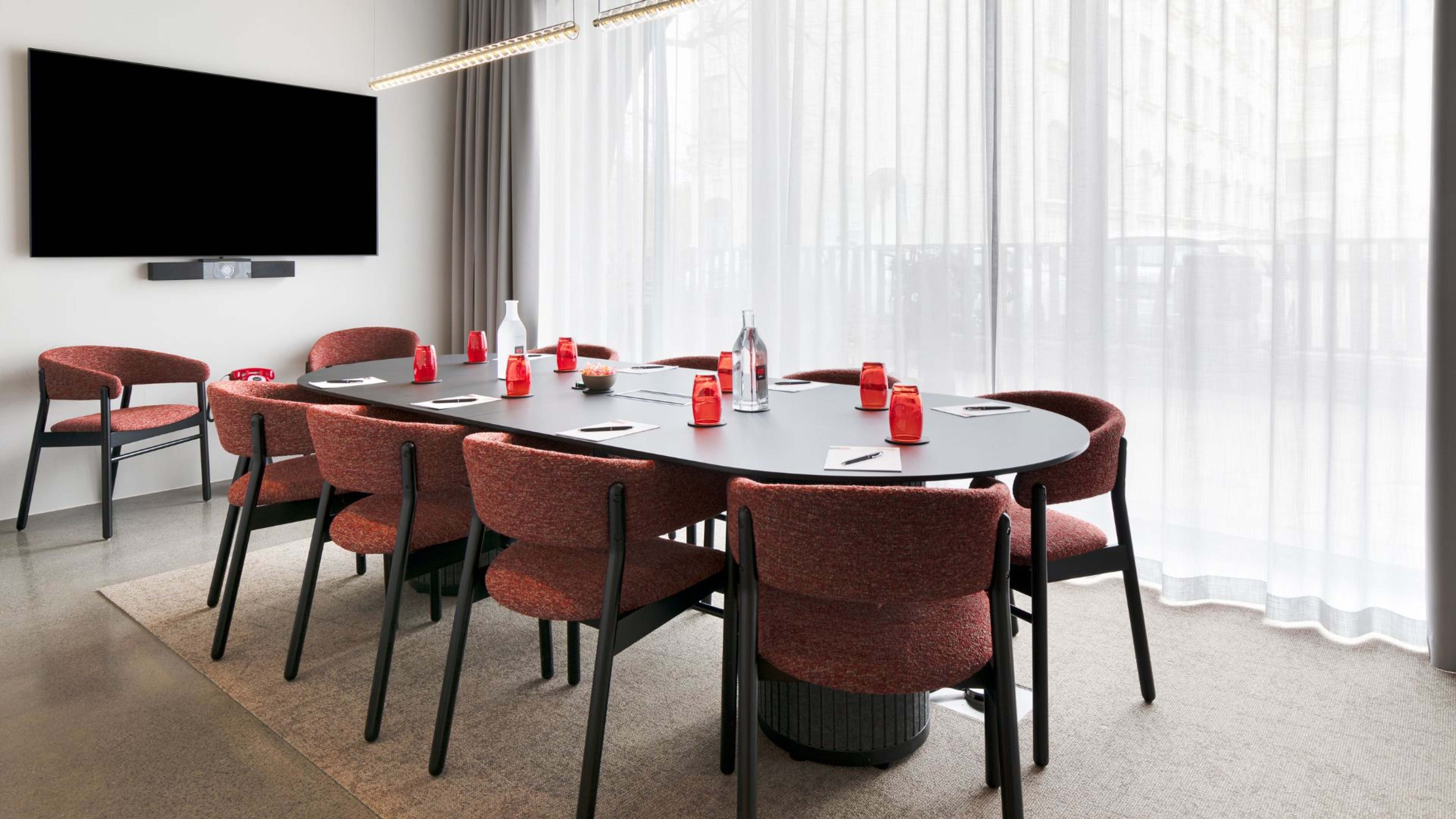 Moderni kokoustila, jossa ovaali musta pöytä, punaruskeilla tuoleilla. Seinällä litteä näyttö, huoneessa isot ikkunat ja tasainen kokolattiamatto.