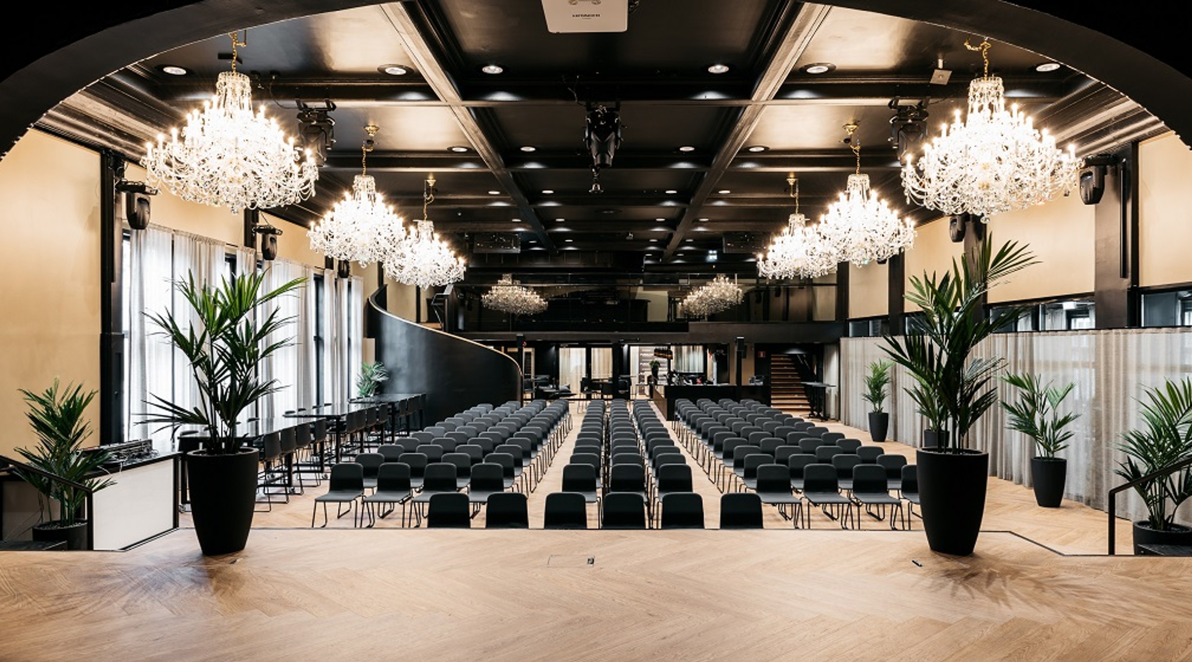 Tyylikäs auditorio kristallikruunuilla, mustilla tuoleilla riveissä ja viherkasveja reunoilla. Lattia on tasainen puulattia.
