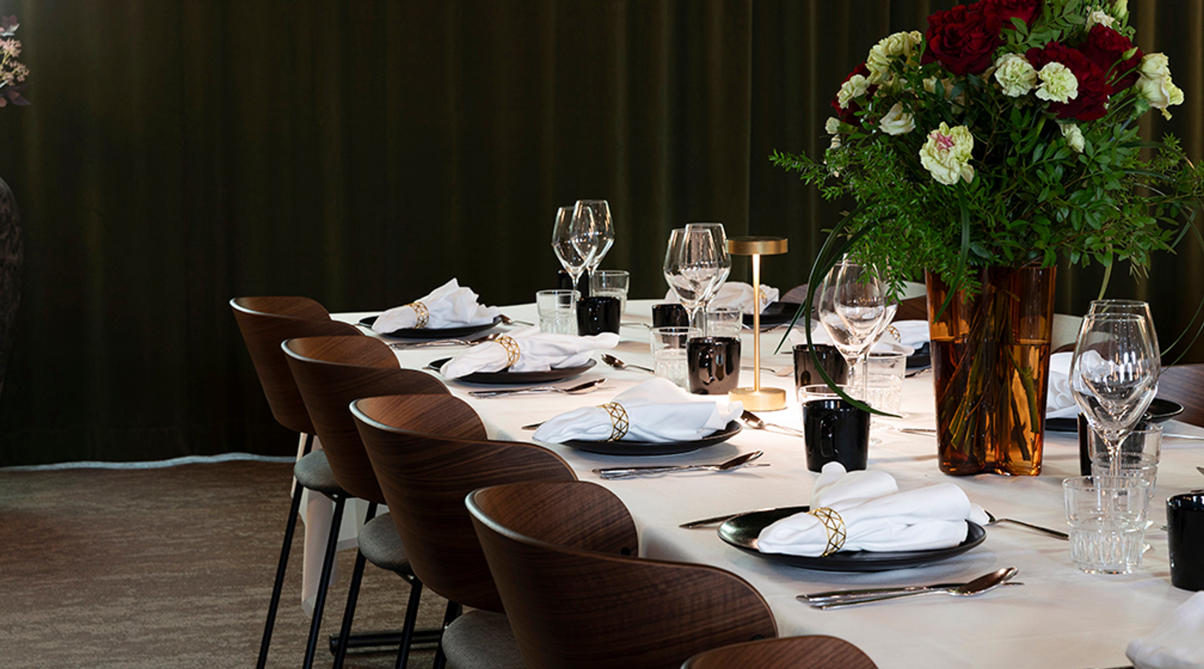 Elegantisti katettu pöytä ruskeilla tuoleilla, valkoisilla lautasliinoilla ja koristeellisella kukka-asetelmalla, jossa punaisia ja valkoisia kukkia.