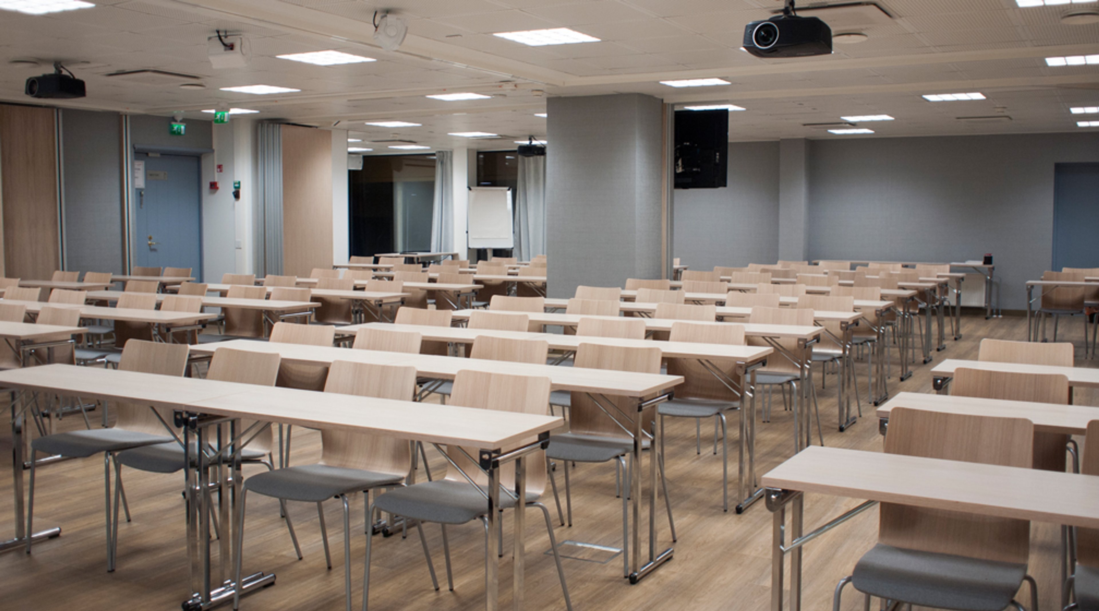 Luokkahuone riveittäin järjestetyillä vaaleilla pöydillä ja tuoleilla, projektorit katossa ja esitystaulu edessä. Tilassa puulattia ja hyvä valaistus.