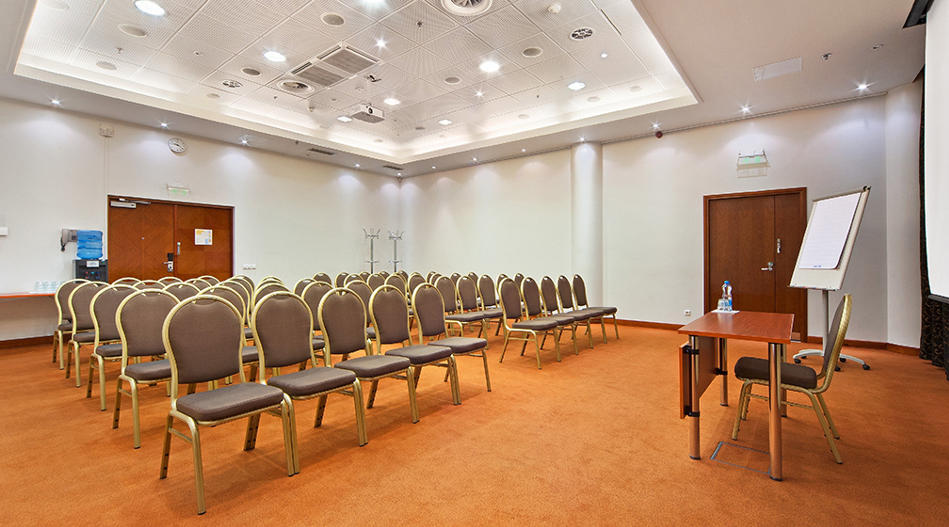 Kokoustila oranssilla matolla ja riveittäin aseteltuja tuoleja. Edessä pöytä, fläppitaulu ja valkokangas. Tilassa kaksi puuovea ja tasainen lattia.