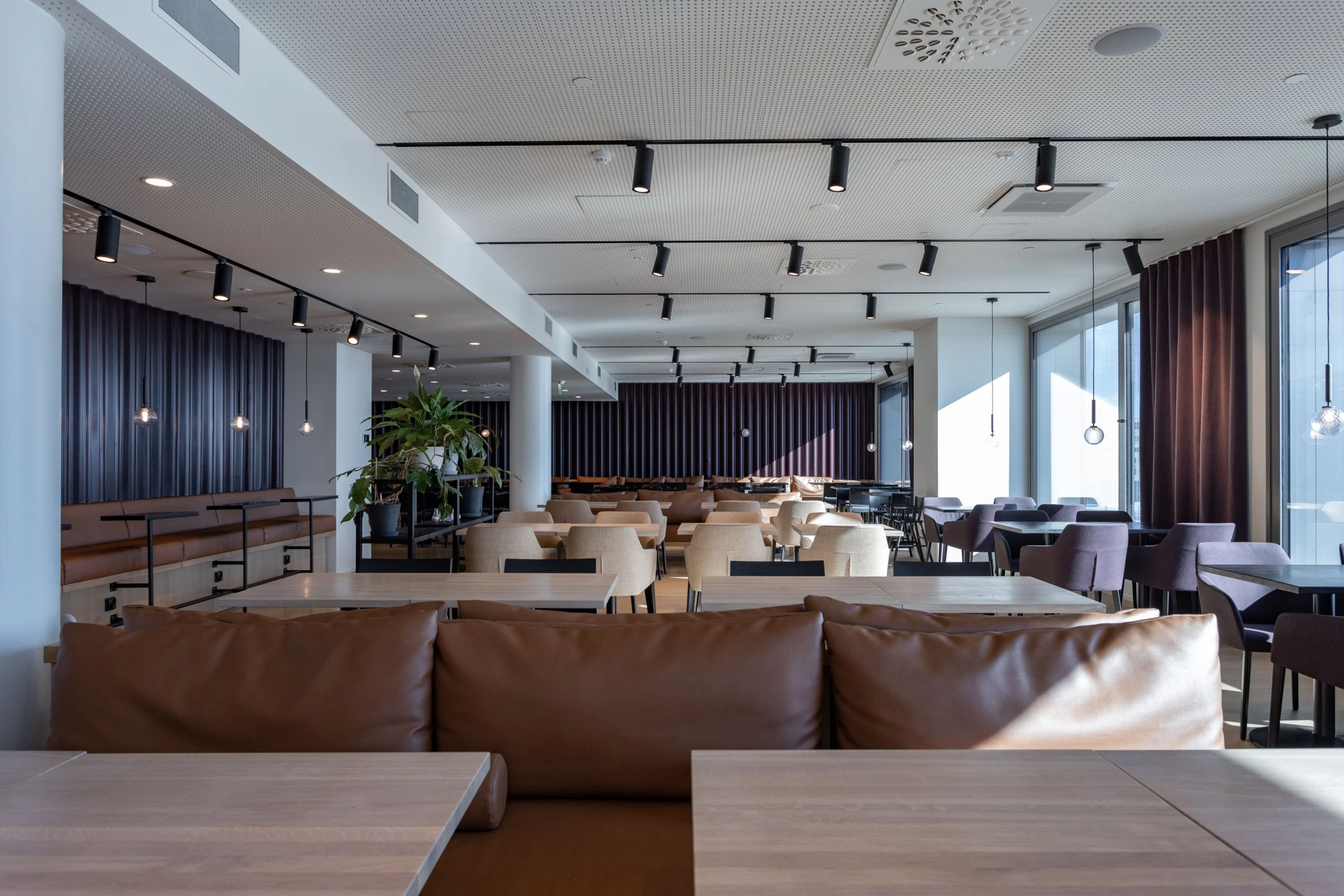 Moderni ravintolasisustus, jossa ruskeita nahkasohvia, vaaleita pöytiä, lattia-ikkunat ja viherkasveja. Tasainen lattia, avara tila.