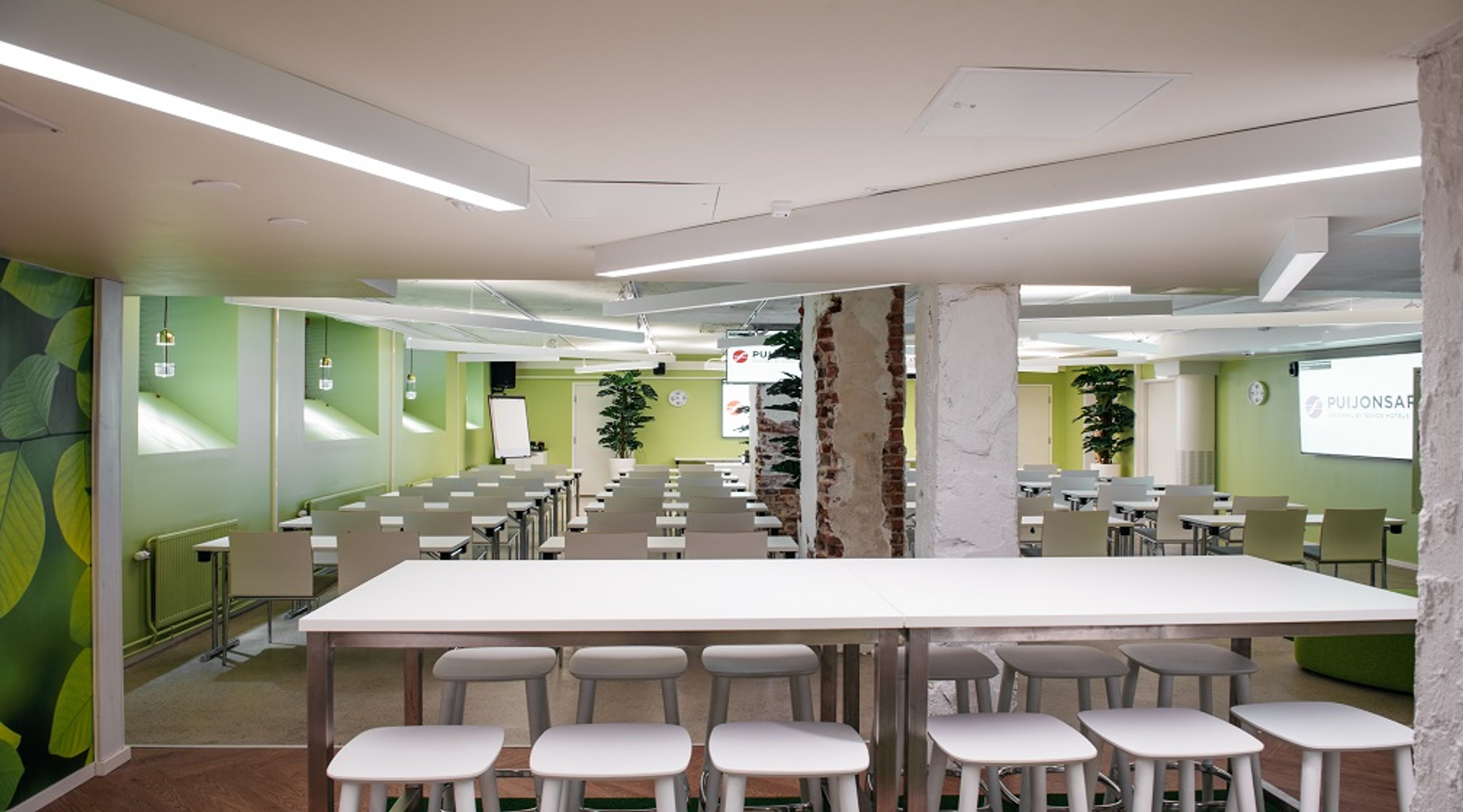 Valoisa kokoustila vihrein seinin ja valkoisin pöydin sekä tuolein. Tila on avara, hyvin valaistu ja sisältää esitystaulun.