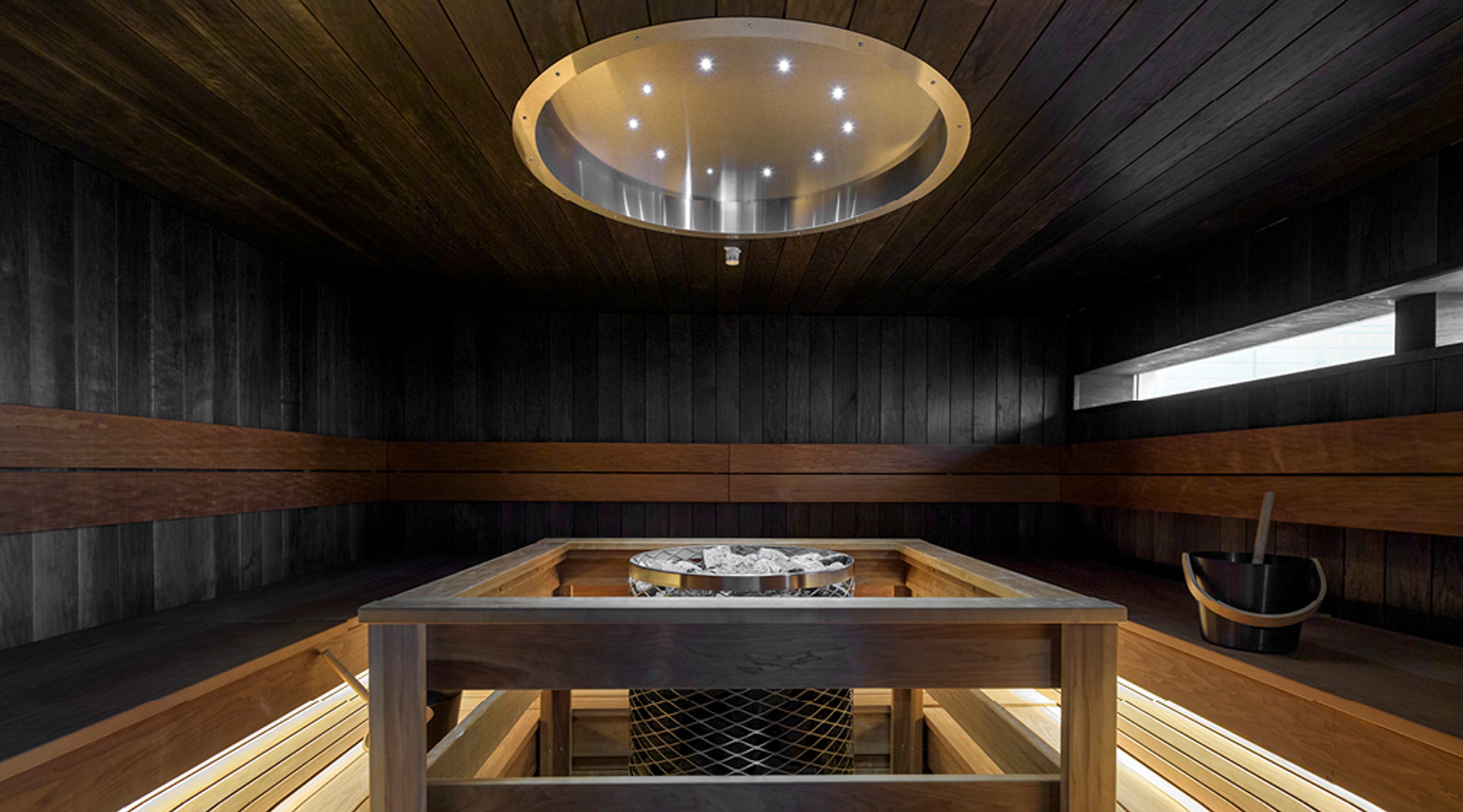 Moderni tumma sauna puupaneloinnilla, pyöreä kattoikkuna, puiset istuintasot ja kiuas keskellä.