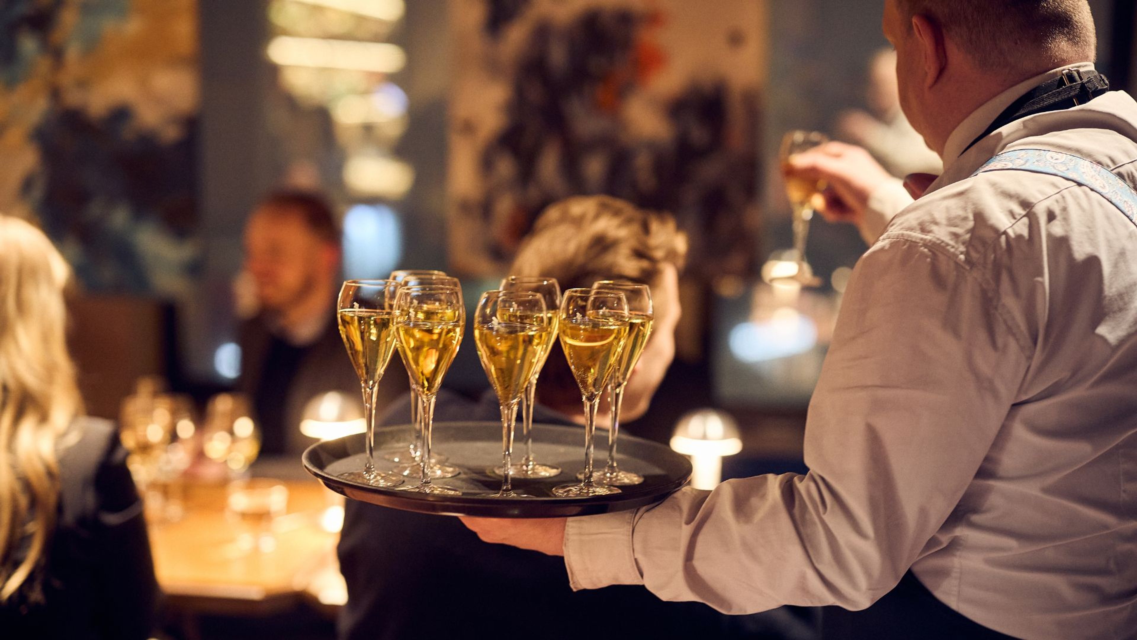 Tarjoilija kantaa tarjottimella täytettyjä samppanjalaseja hämärässä ravintolassa.