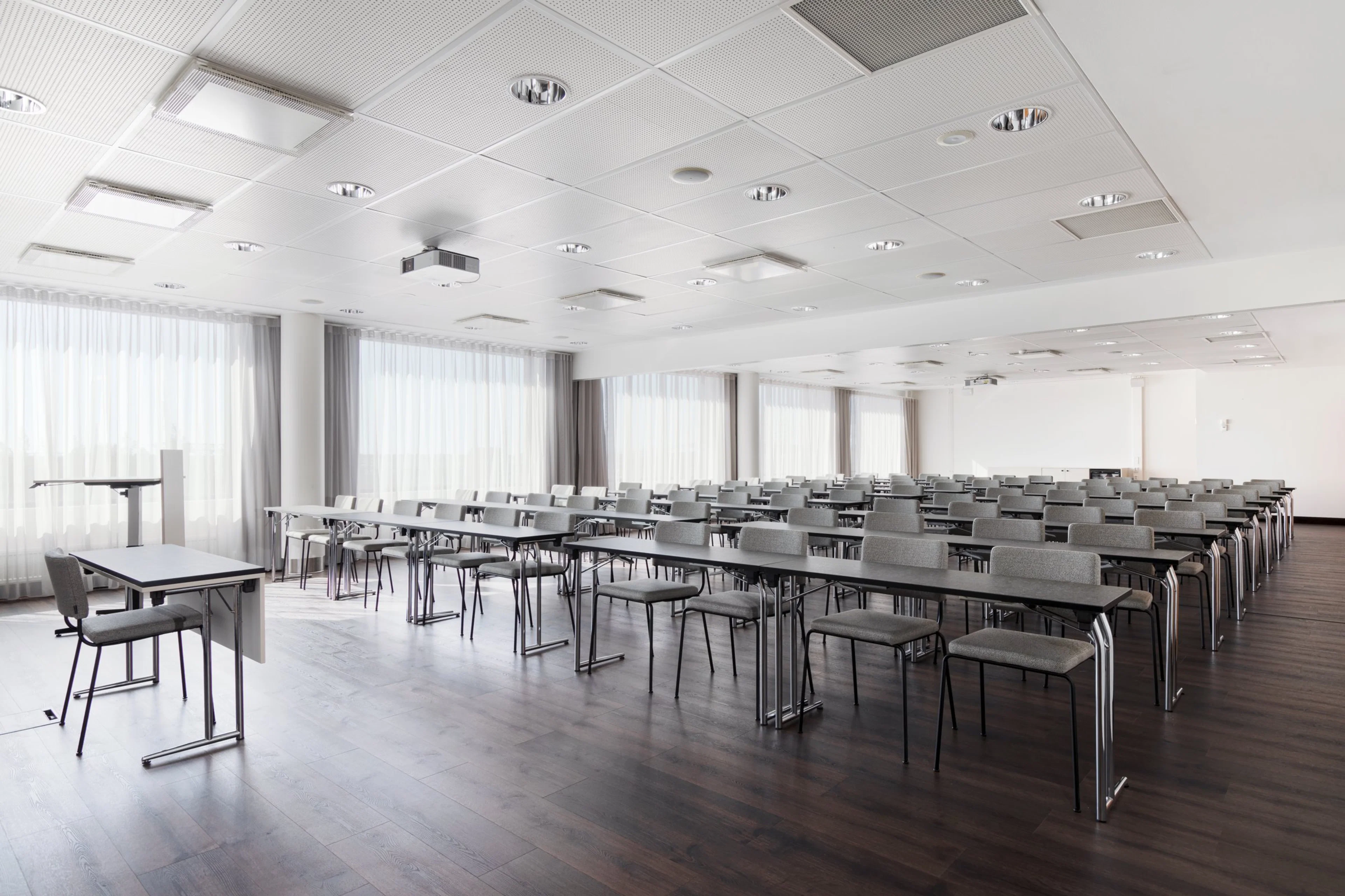 Moderni kokoustila useilla pöydillä ja tuoleilla riveissä. Valkoinen sisustus, puulattia ja suuret ikkunat valkoisilla verhoilla. Puhujapönttö etualalla.