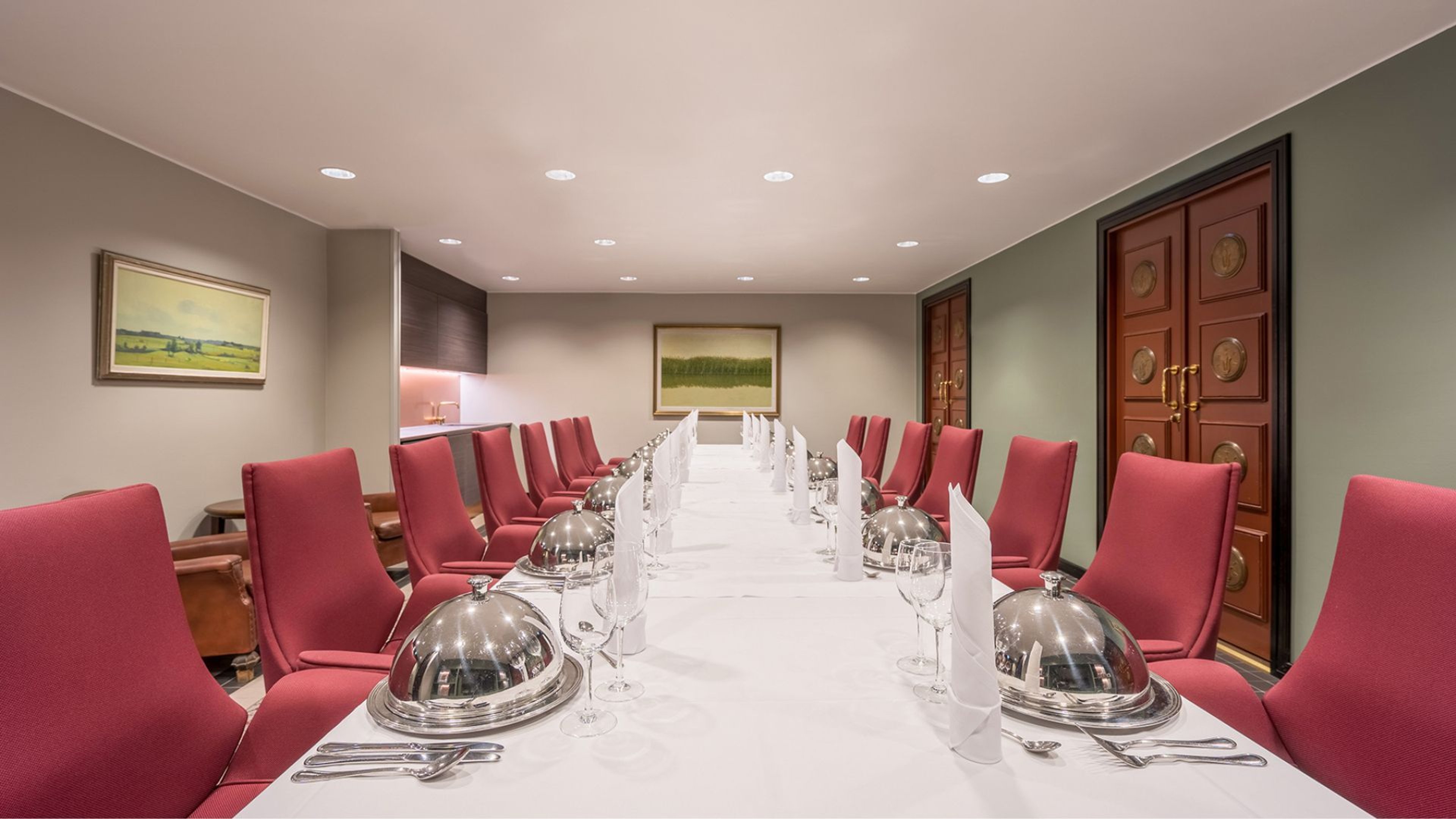 Elegantti kokous- tai juhlatila, jossa pitkä valkoinen pöytä, punaisia tuoleja, hopeakupuja ja maisematauluja seinällä. Tila on hyvin valaistu.