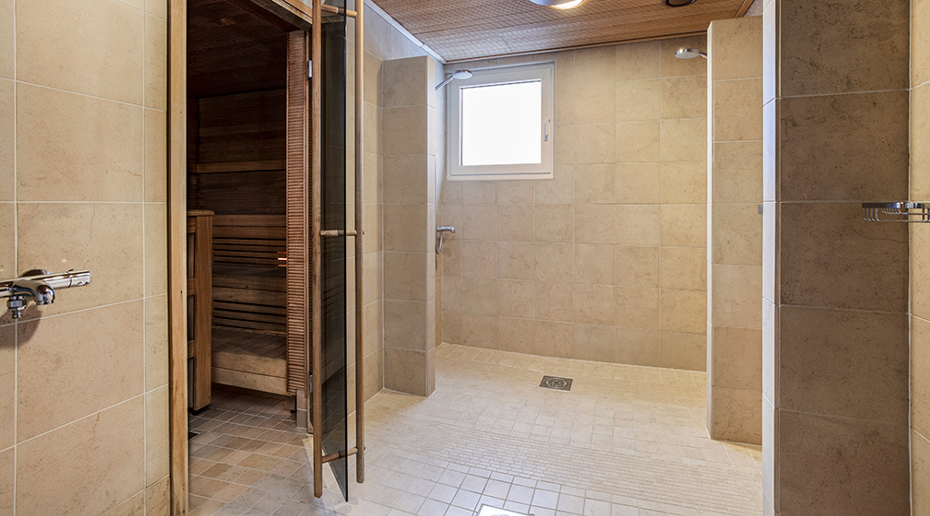 Moderni saunatilojen yhdistelmä, jossa avoimella ovella varustettu puinen sauna ja vieressä beige-laatoitettu suihkutila ikkunalla.