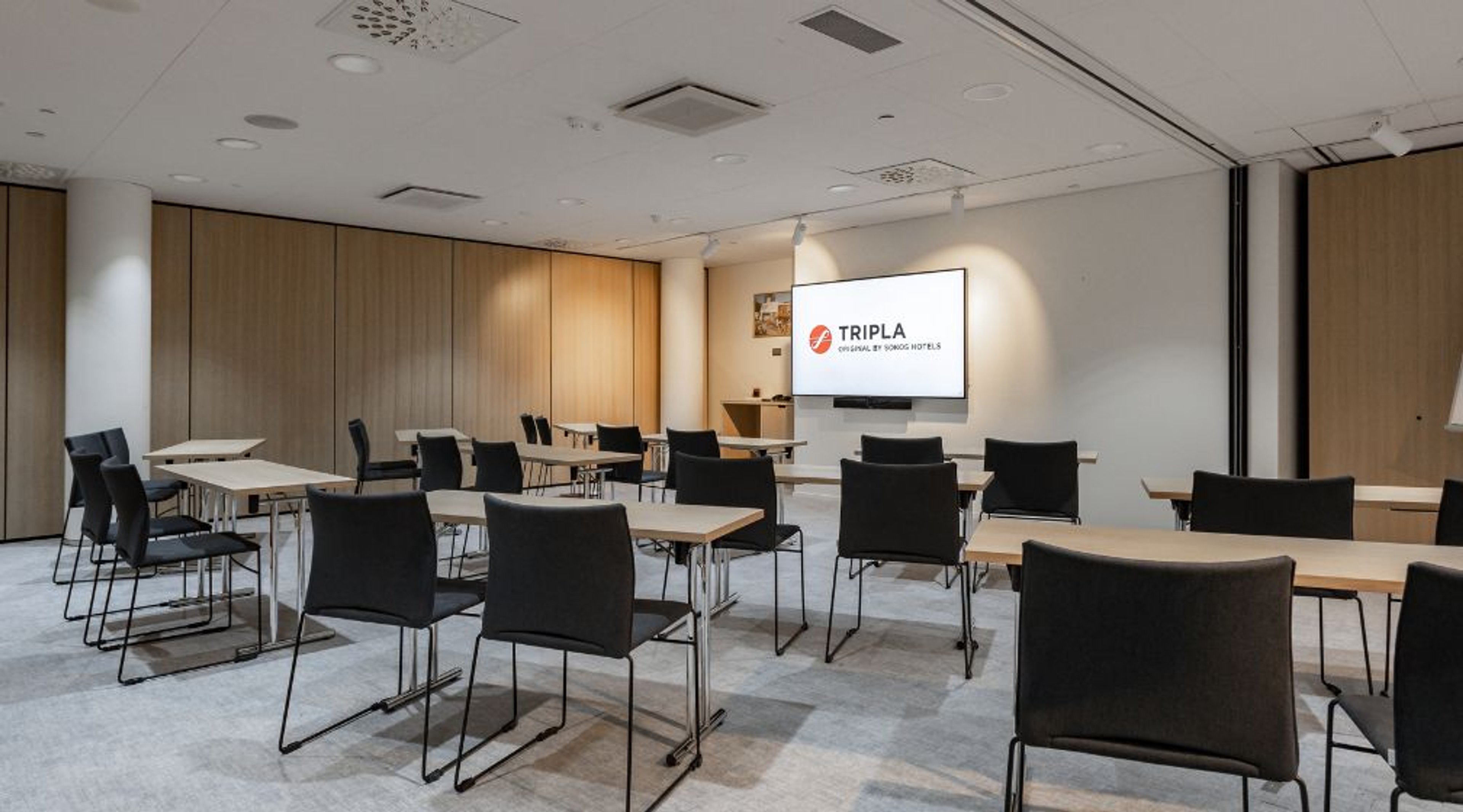 Moderni kokoustila, jossa kevyitä pöytiä ja tuoleja luokkamuodostelmassa. Edessä iso näyttö ja puiset seinäpaneelit. Vaalea lattia ja hyvä valaistus.