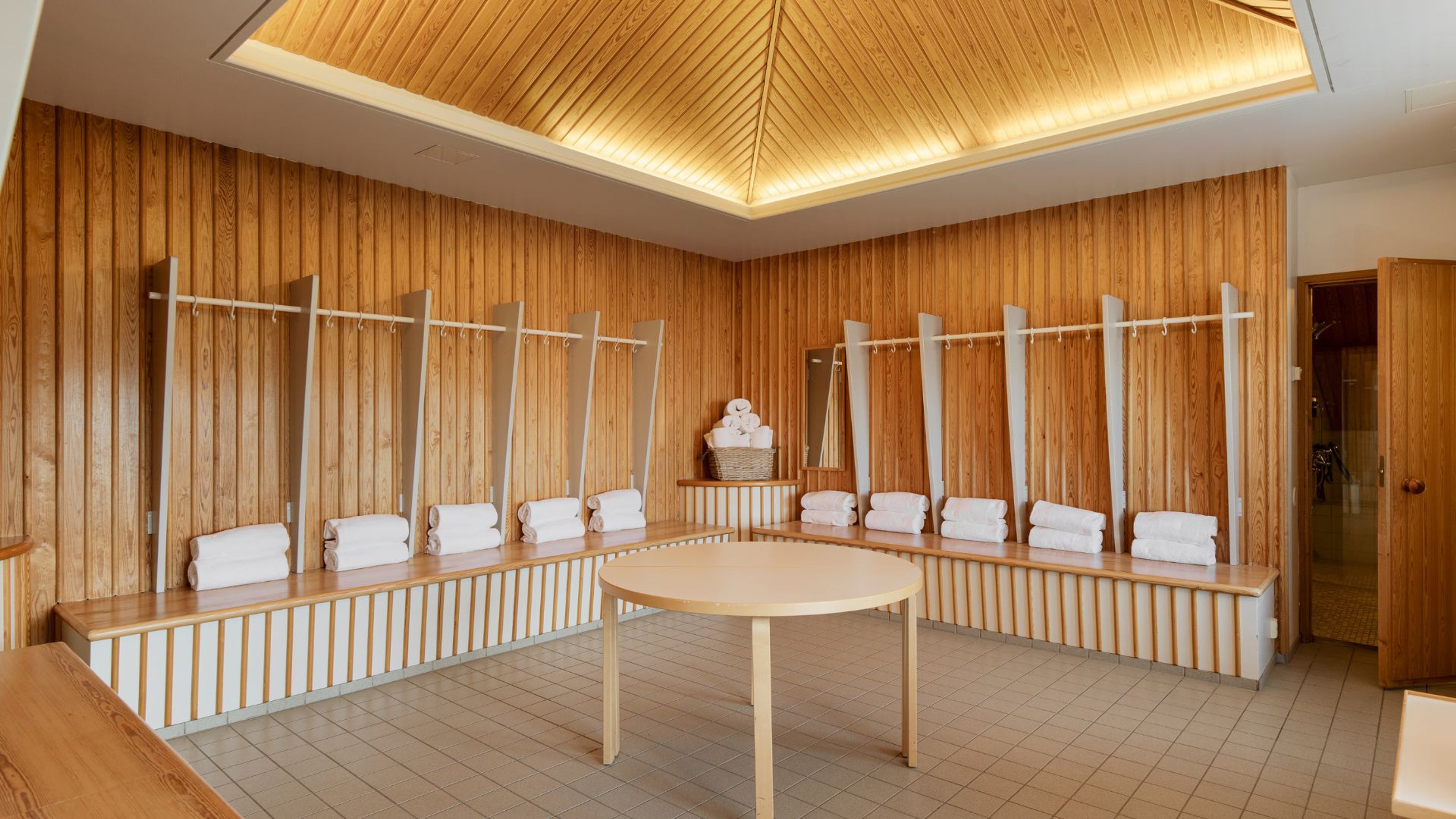 Puupaneloitu saunan pukuhuone, jossa pitkät penkit, valkoisia pyyhkeitä, pyöreä pöytä ja epäsuora kattovalaistus.