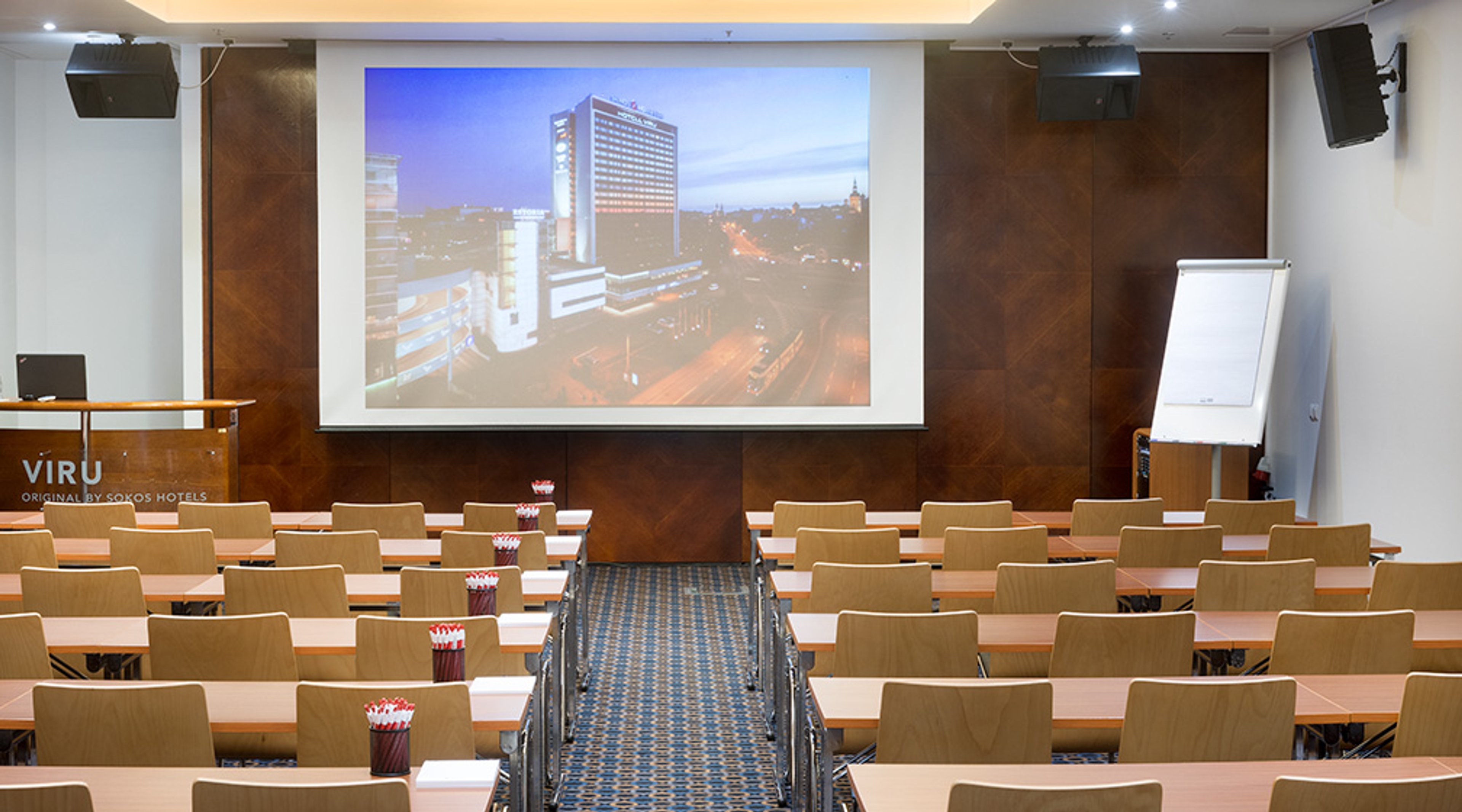 Konferenssihuone riveittäin järjestetyillä puisilla pöydillä ja tuoleilla, keskellä sininen käytävä ja edessä suuri projektori. Seinillä puupaneelit.