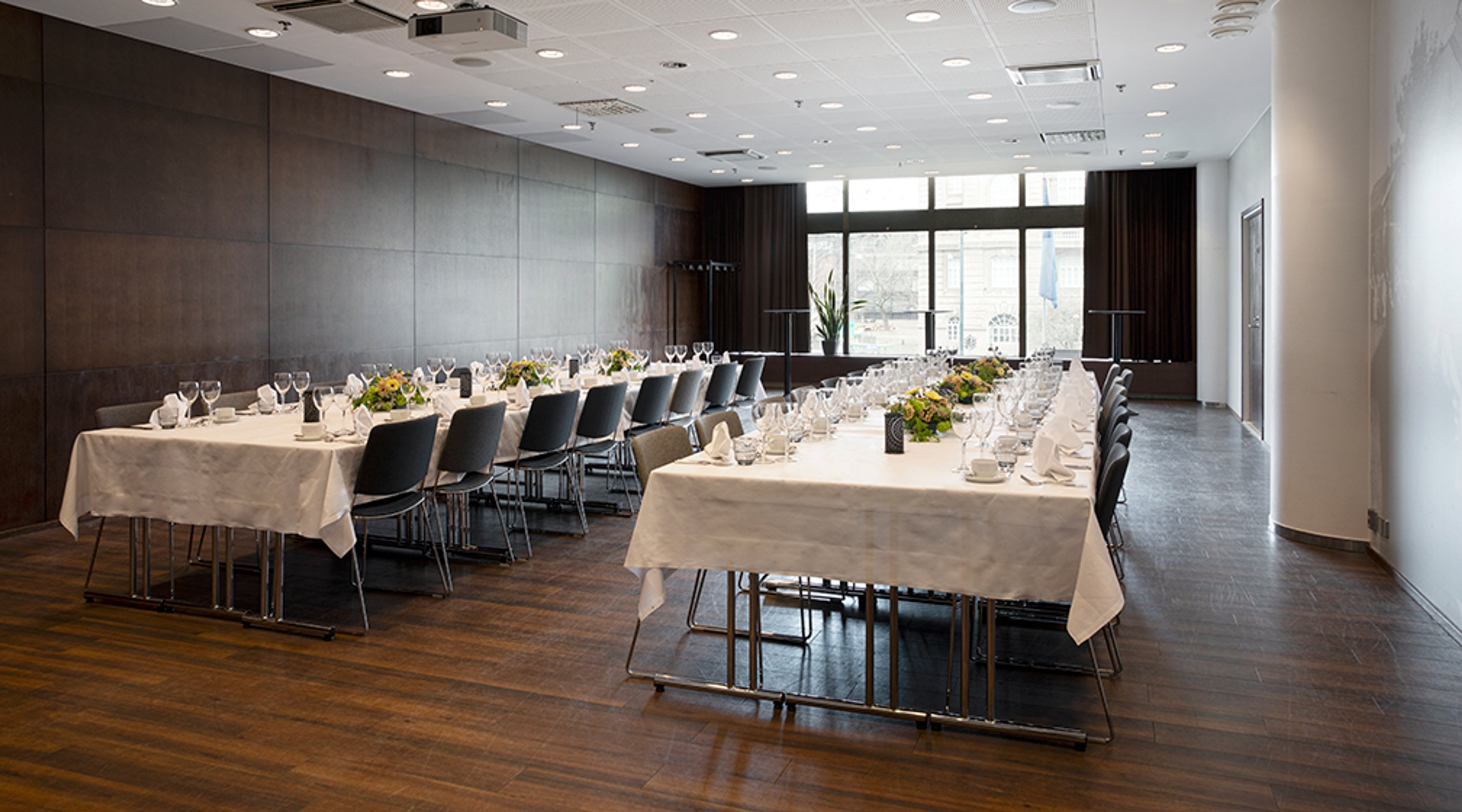 Kokoustila, jossa on kaksi pitkää valkoliinalla katettua pöytää. Tumma puulattia, ruskeat seinäpaneelit ja isot ikkunat. Hyvä valaistus.