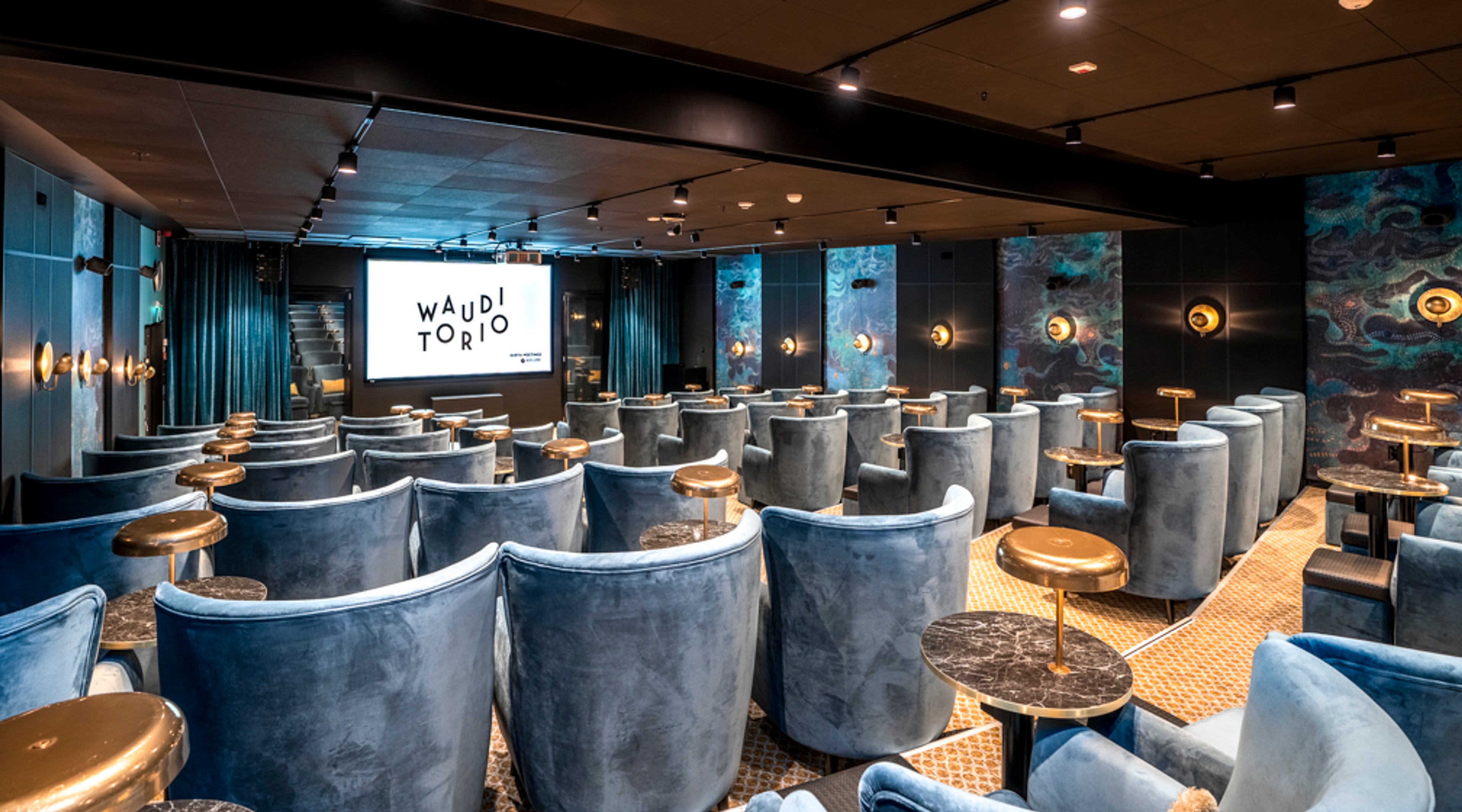 Moderni auditorio sinisillä nojatuoleilla ja pyöreillä sivupöydillä. Seinillä koristeellisia paneeleita ja valaistus on himmeä.