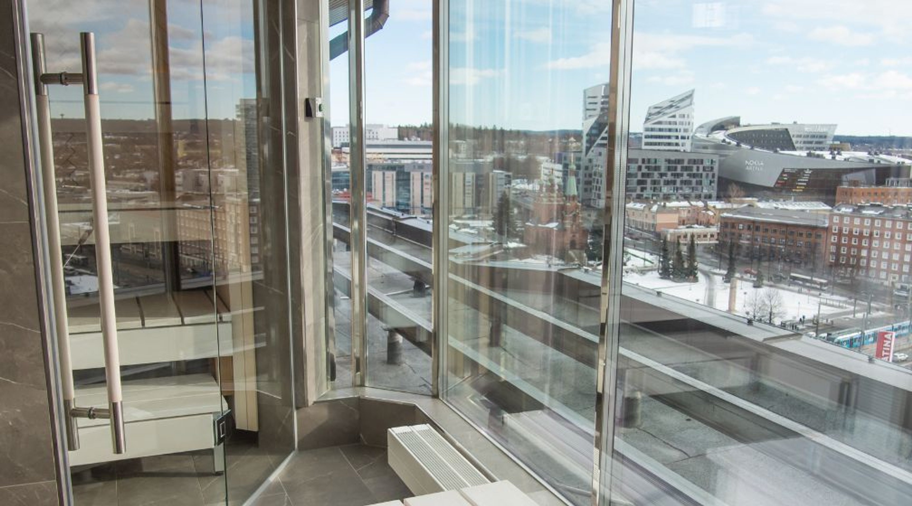 Panoraamanäkymä korkeasta rakennuksesta talviseen kaupunkimaisemaan. Lasitetulta sisäparvekkeelta näkyy moderneja rakennuksia ja luminen ympäristö.