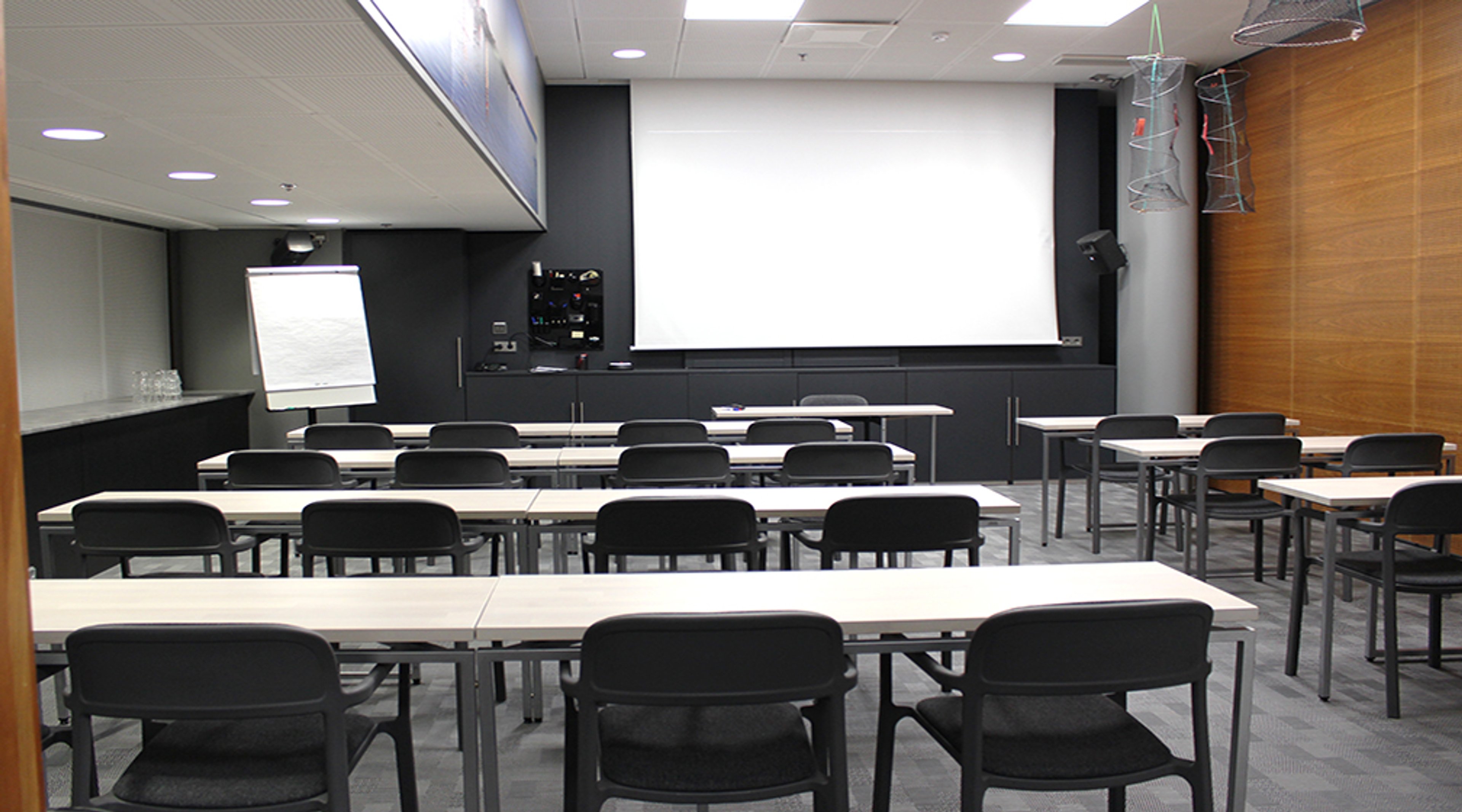 Koulutustila, jossa on valkoisia pöytiä ja mustia tuoleja riveittäin, edessä valkokangas ja fläppitaulu. Huoneessa puupaneeli seinä ja tasainen valaistus.