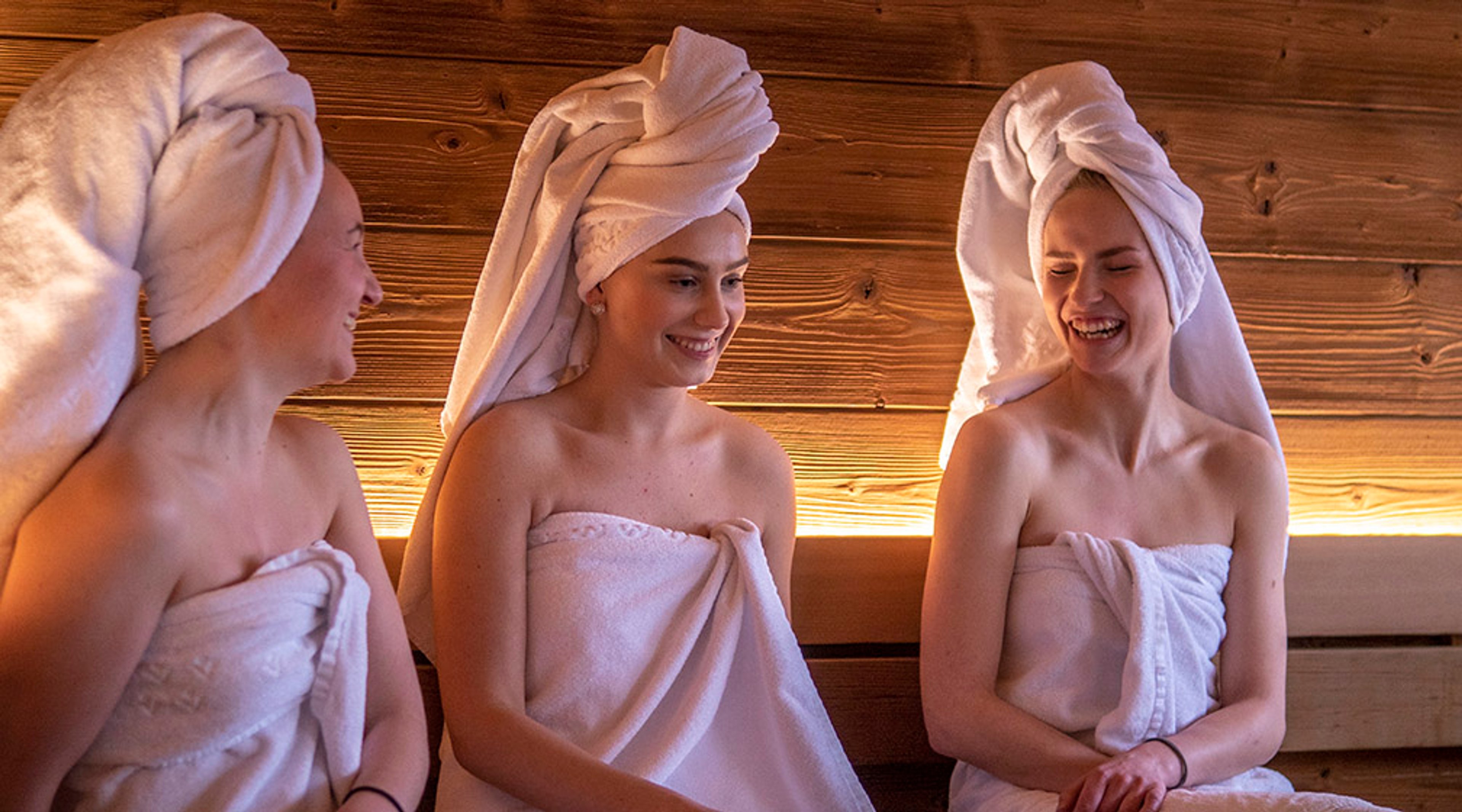 Kolme henkilöä saunassa tai spa-tilassa, kietoutuneina valkoisiin pyyhkeisiin, hiukset pyyhkeellä suojattuna, puupaneeliseinä taustalla.