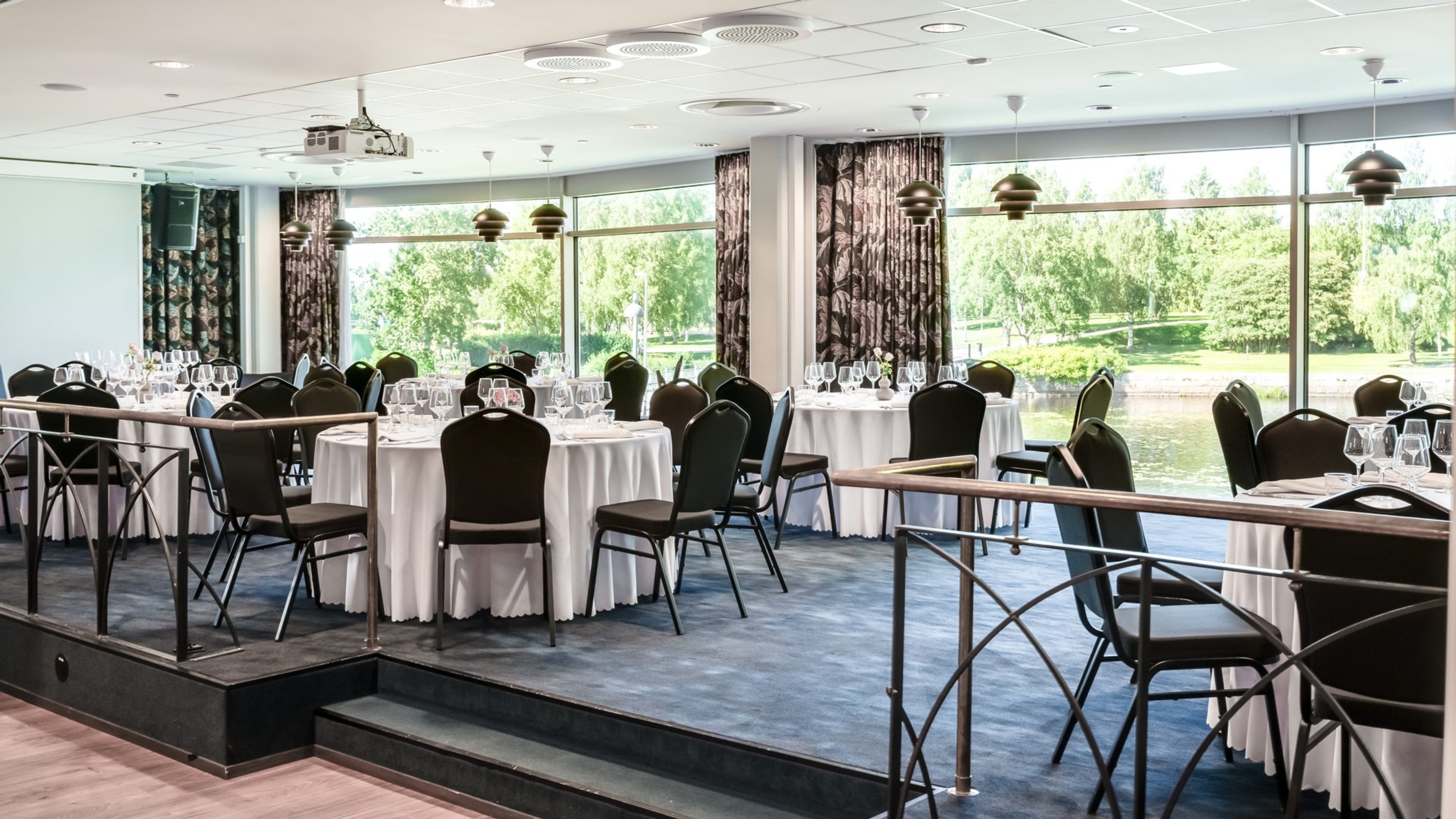 Ravintolasali, jossa valkoisia pöytiä ja mustia tuoleja. Tilassa on korotettu lattia ja suuret ikkunat, joista näkyy puistomainen maisema ja vesistö.