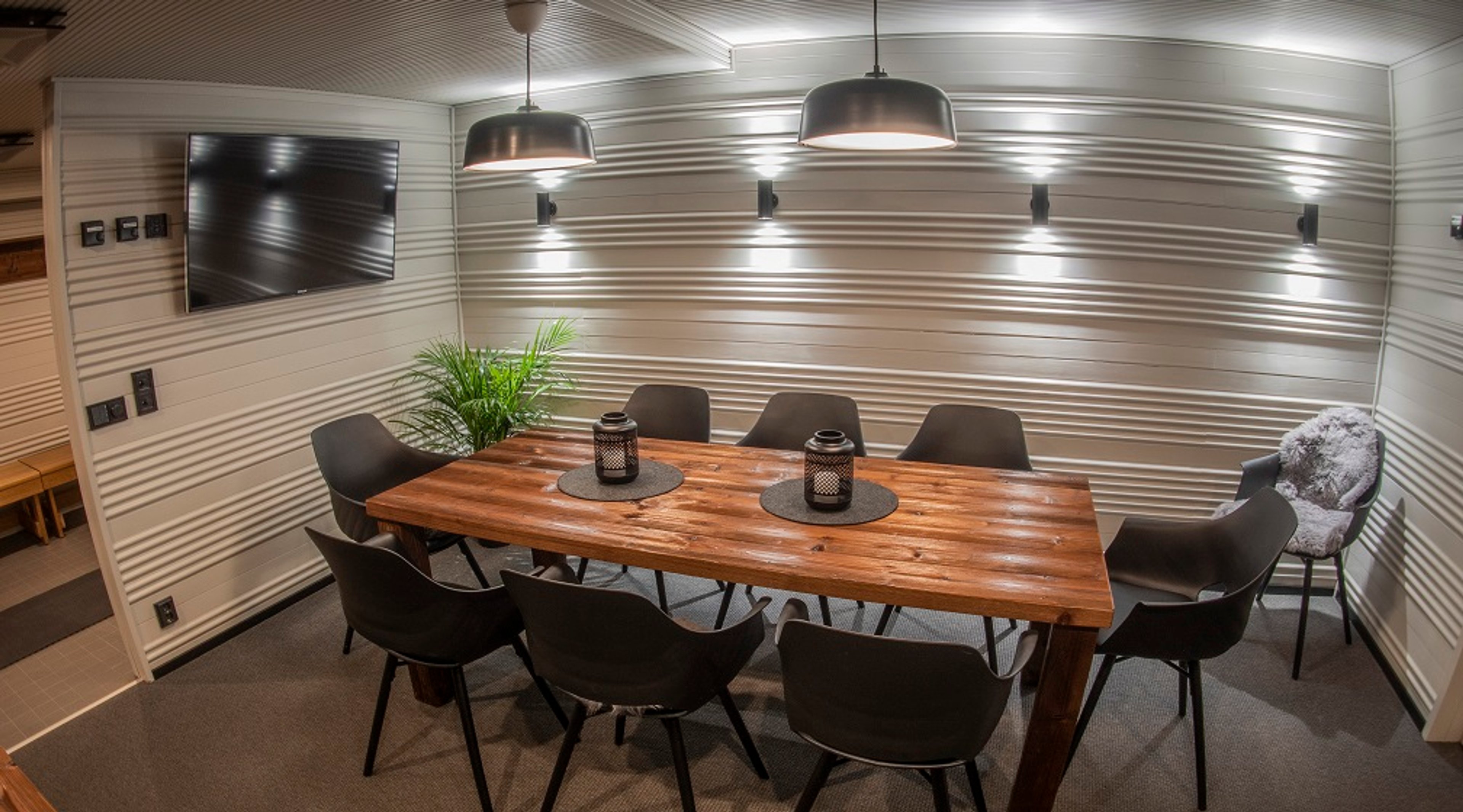 Moderni neuvotteluhuone, jossa puinen pöytä, mustat tuolit, TV-näyttö seinällä ja hyvä valaistus. Huoneessa on matto lattialla ja viherkasvi.