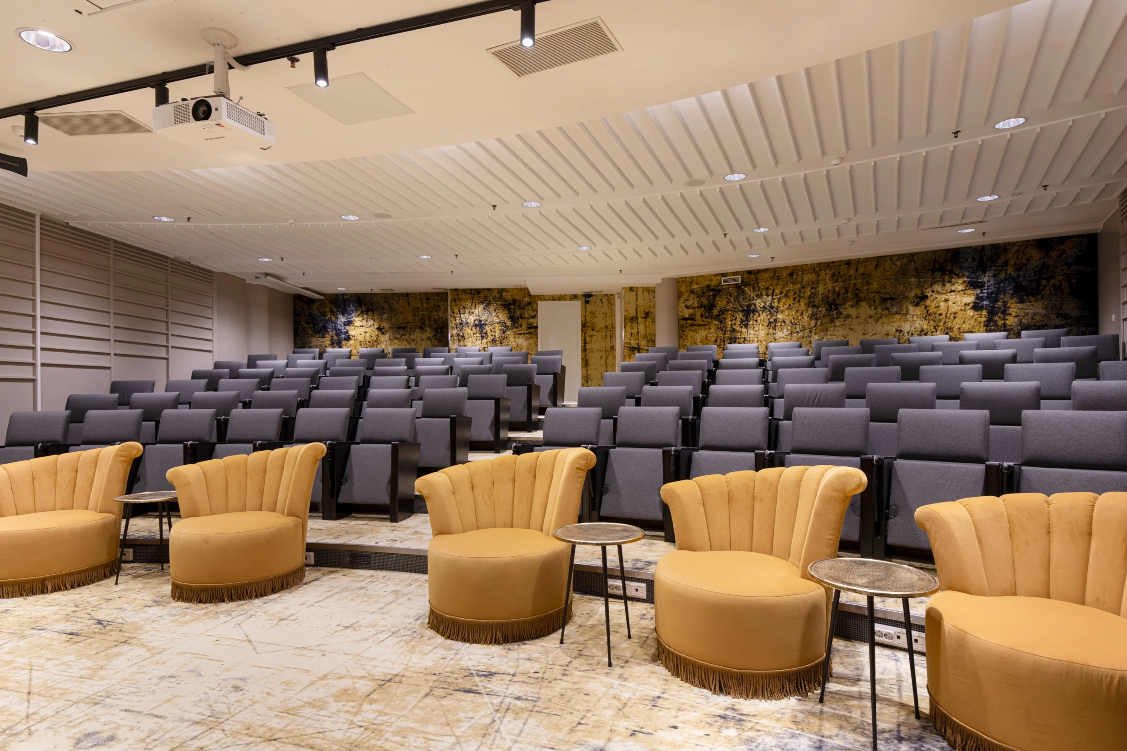 Auditorium with rows of gray seats and yellow armchairs in the front row. Features marble flooring and a stylish gold back wall.