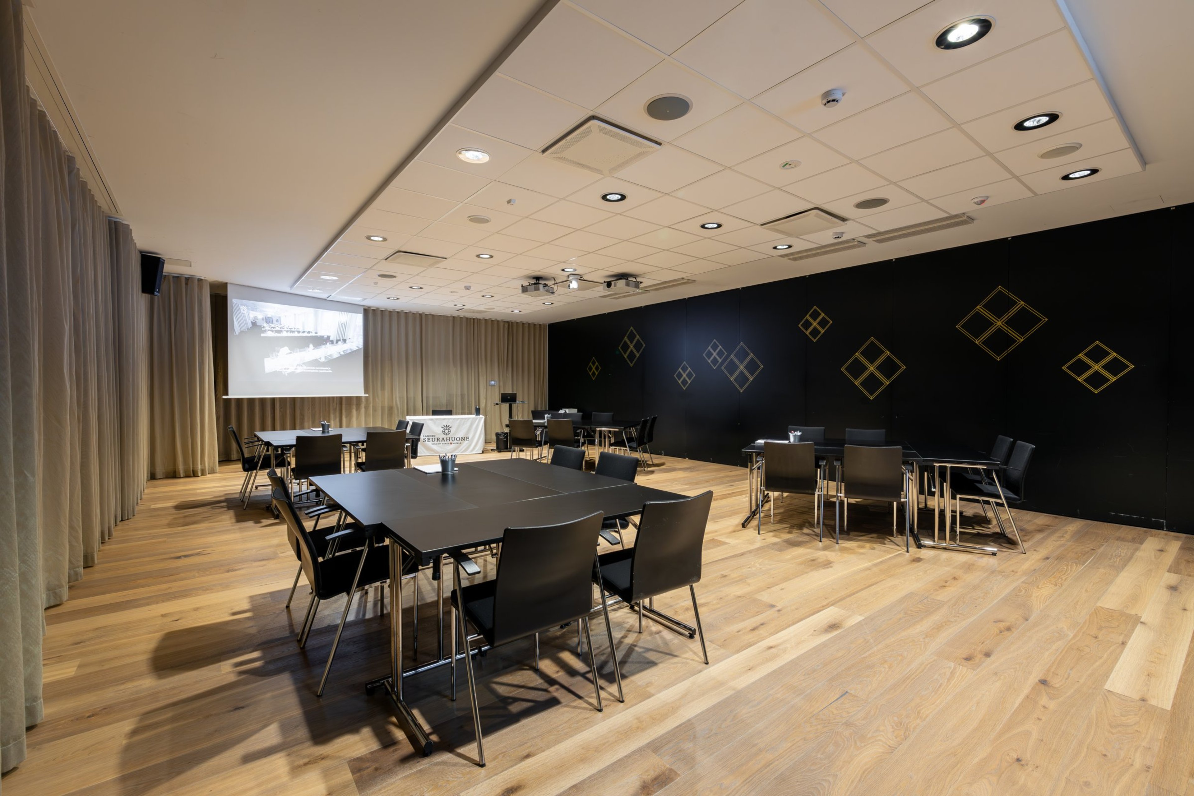 Nykyaikainen kokoustila puulattialla. Mustia pöytiä ja tuoleja, musta takaseinä koristeilla, vaalea katto, projektori ja verhot seinillä.