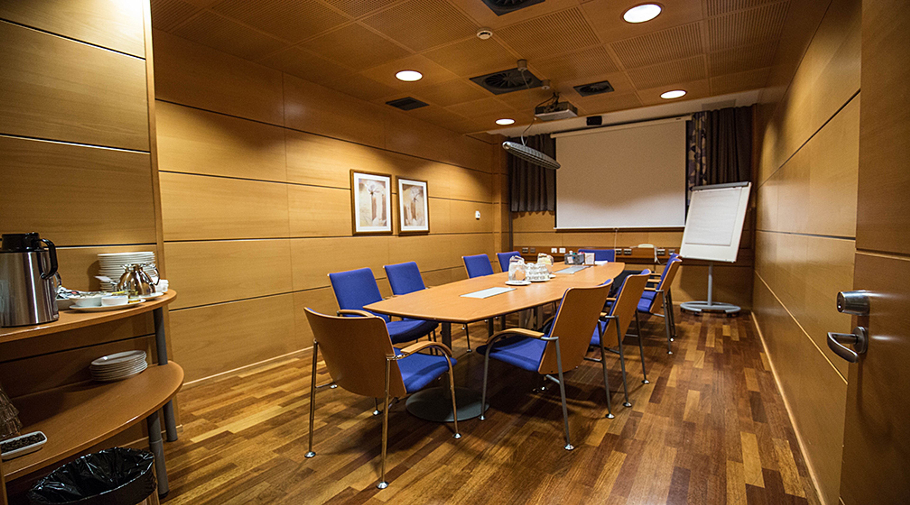 Kokoustila puupaneeliseinillä, pitkä pöytä sinisillä tuoleilla, projektori ja valkotaulu. Puulattia ja tarjoilupöytä.