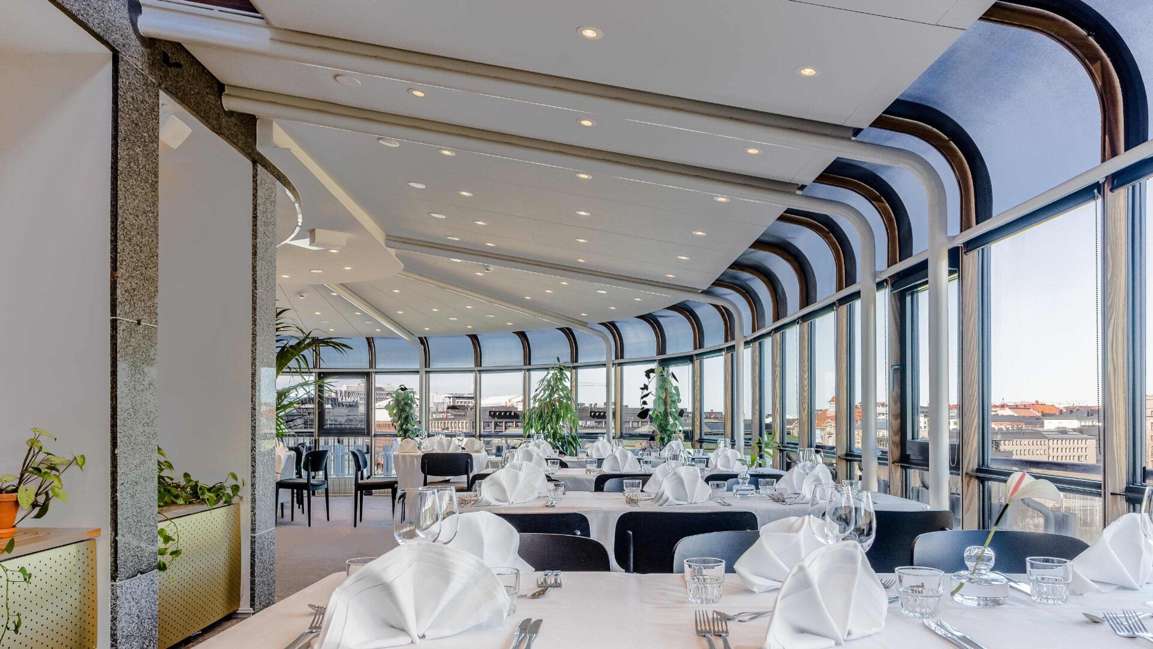 Kaarevassa panoraamaravintolassa on valkoisia pöytiä ja mustia tuoleja. Korkeat ikkunat tarjoavat näköalan kaupunkiin. Lattia on tasainen, valaistus hyvä.