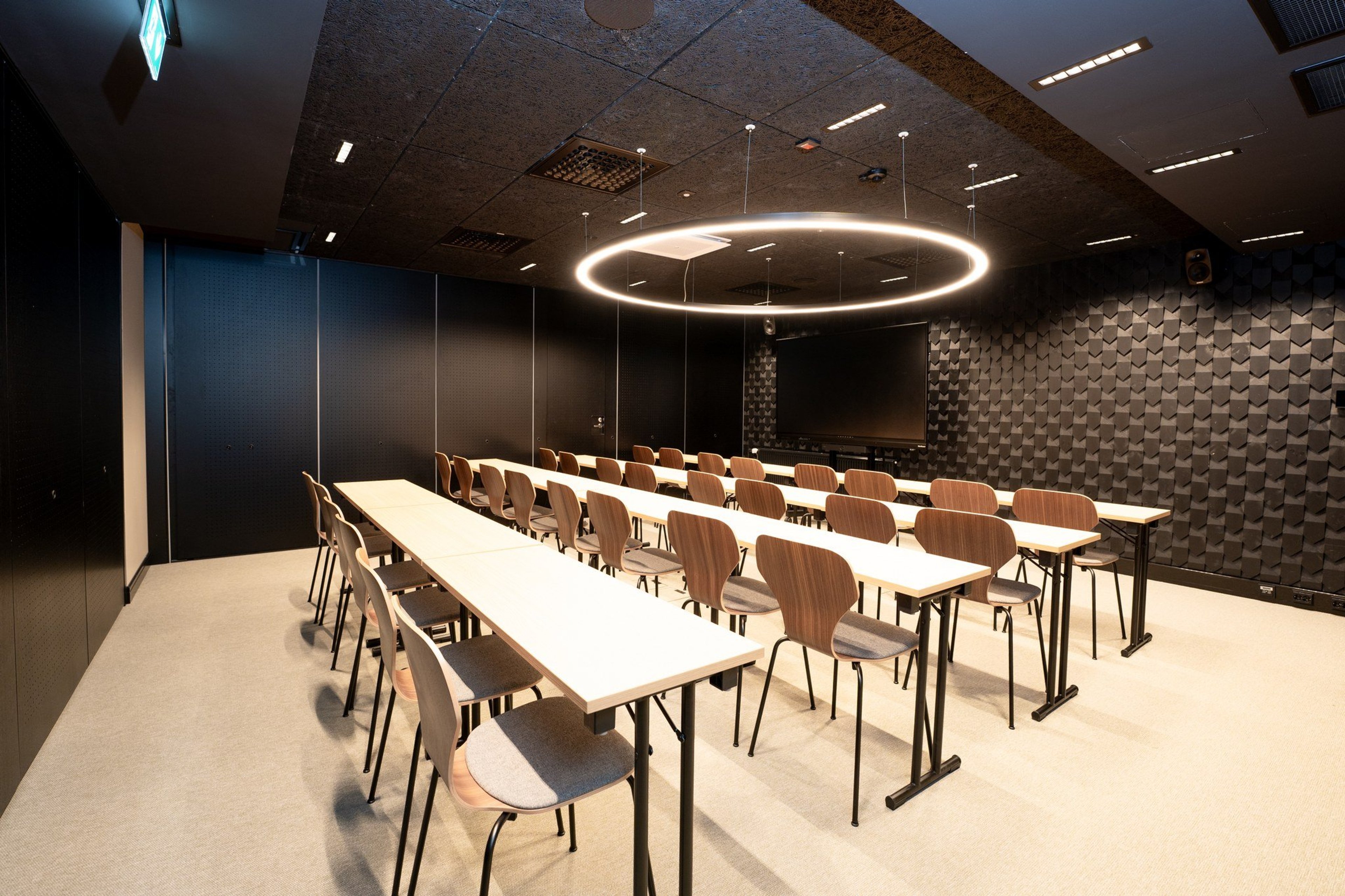 Moderni neuvotteluhuone, jossa vaaleat pöydät ja puiset tuolit. Katossa pyöreä valaisin ja seinällä näyttö. Tilassa tasainen lattia ja hyvä valaistus.