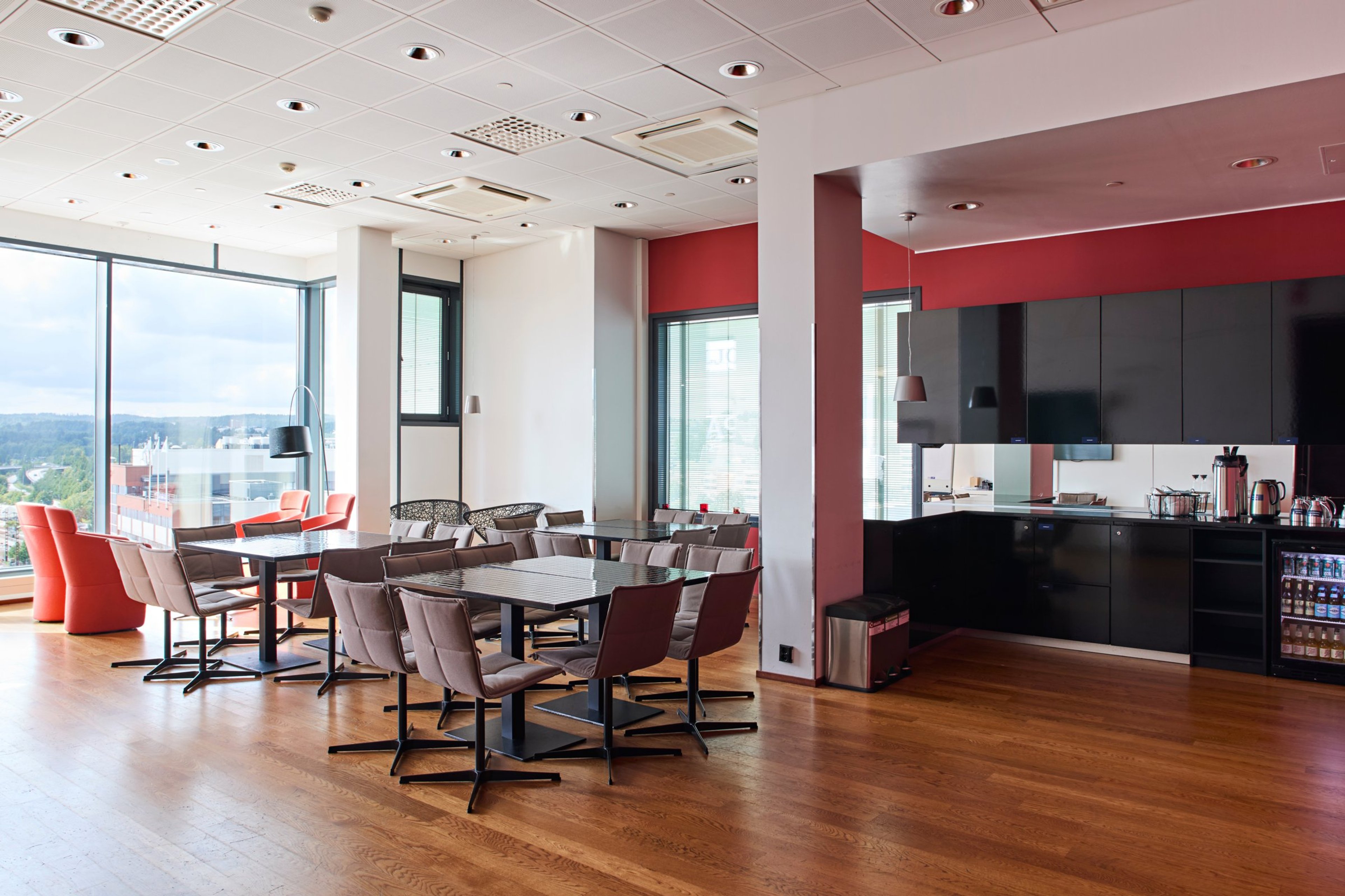 Moderni taukotila, jossa pöytiä ja tuoleja, avoin keittiö mustilla kalusteilla, punaisilla seinäaksenteilla ja suurilla ikkunoilla kaupunkinäkymiin.