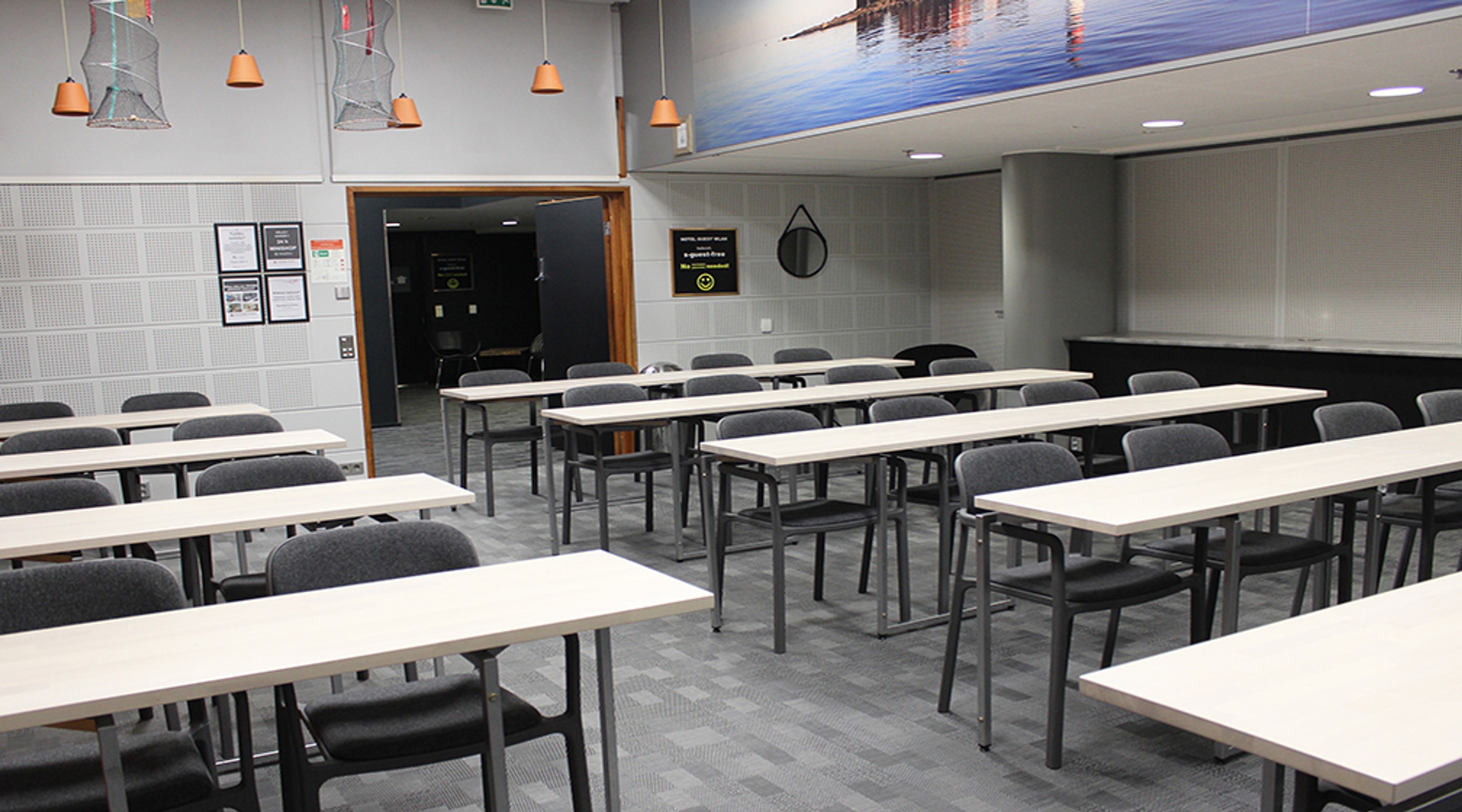 Luokkahuone valkoisilla pöydillä ja harmailla tuoleilla. Laaja sisäänkäynti takaosassa. Sisätilassa hyvä valaistus ja kattopaneelit.