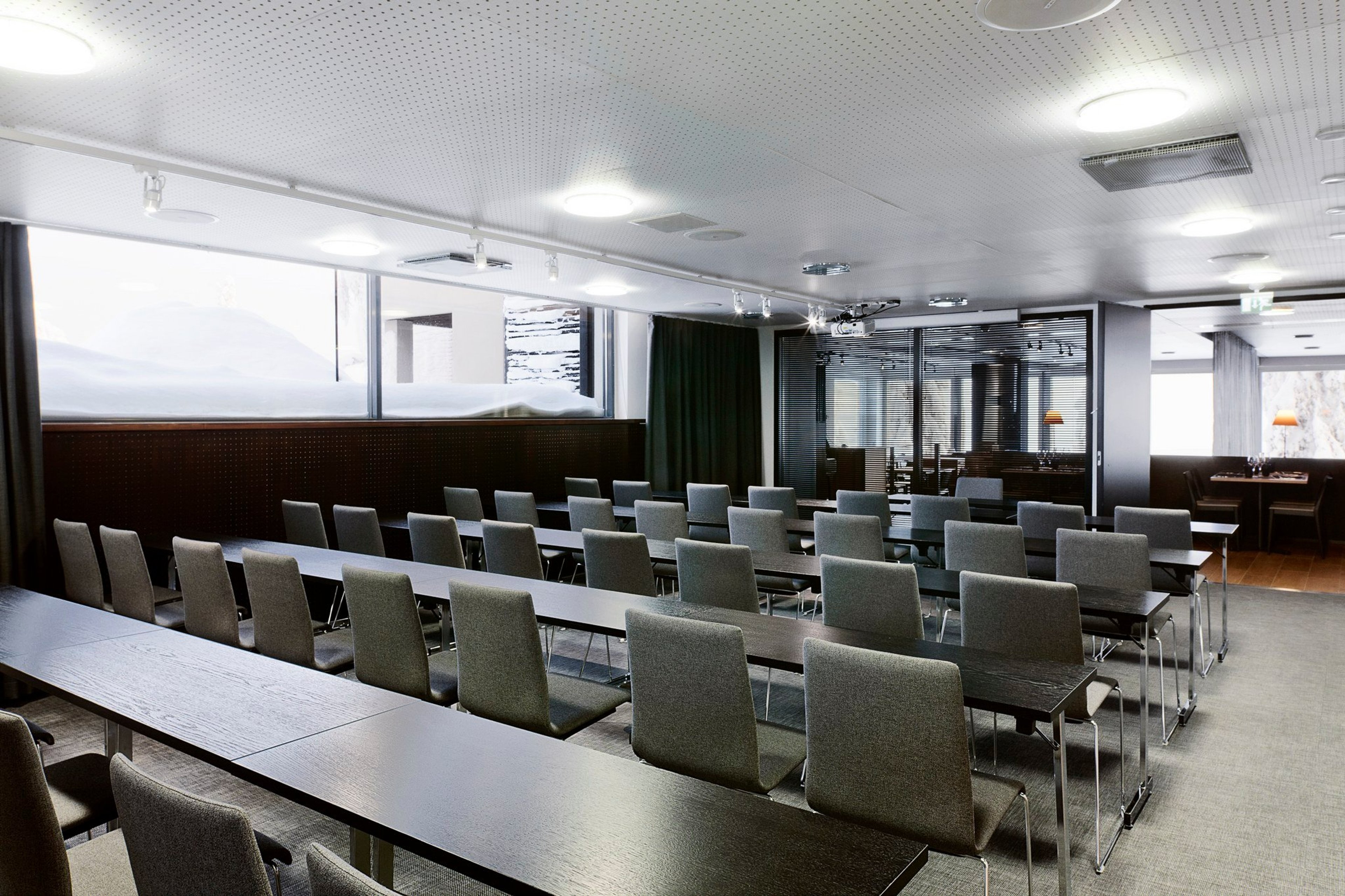 Moderni kokoustila, jossa pitkiä pöytiä ja harmaita tuoleja luokkahuoneasettelussa. Tilassa hyvä valaistus ja isot ikkunat.