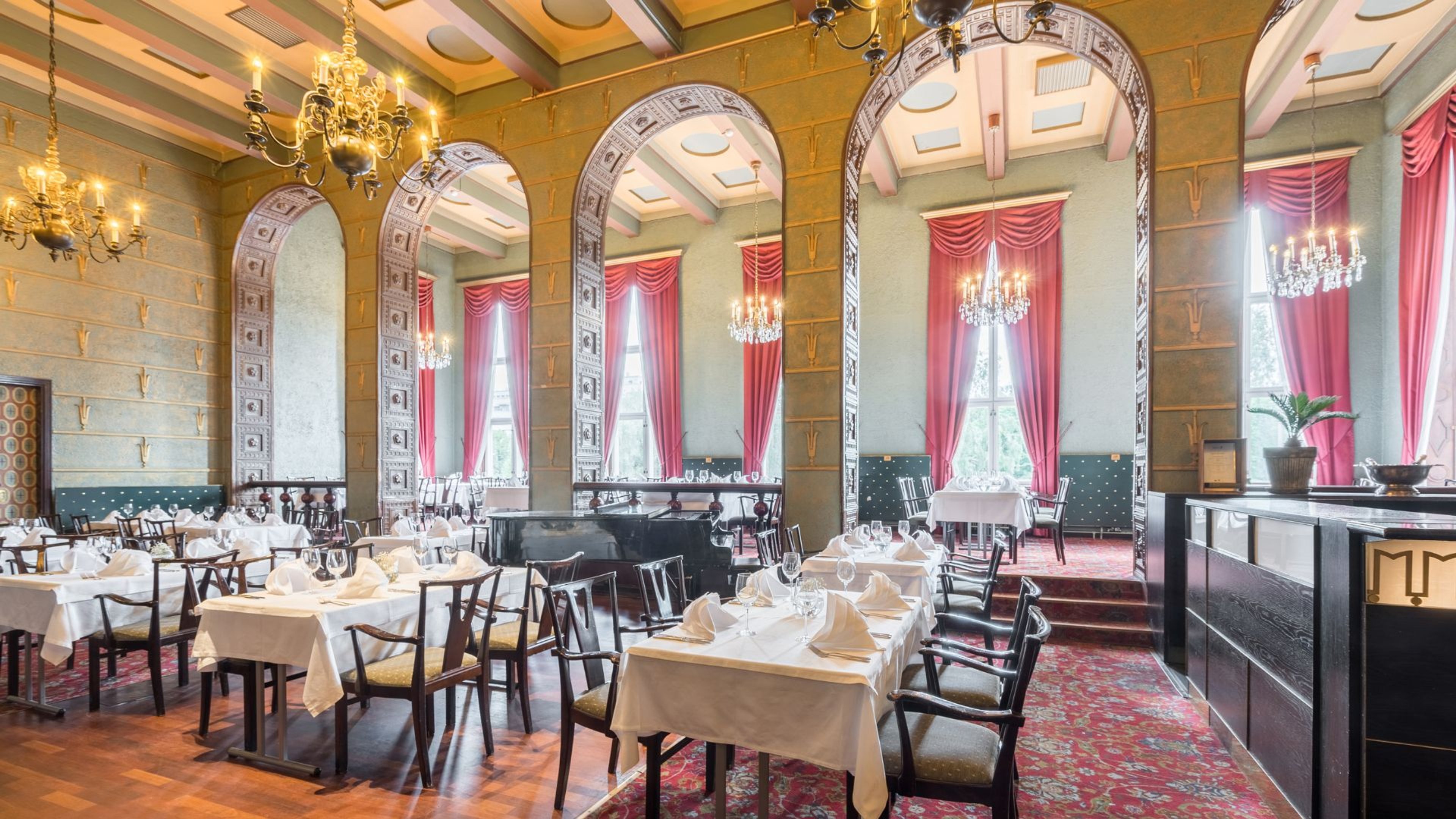 Juhlava ravintolasali, jossa korkeat kaarelliset oviaukot, punaiset verhot, kristallikruunut ja elegantti pöytäkattaus. Tasainen parkettilattia.