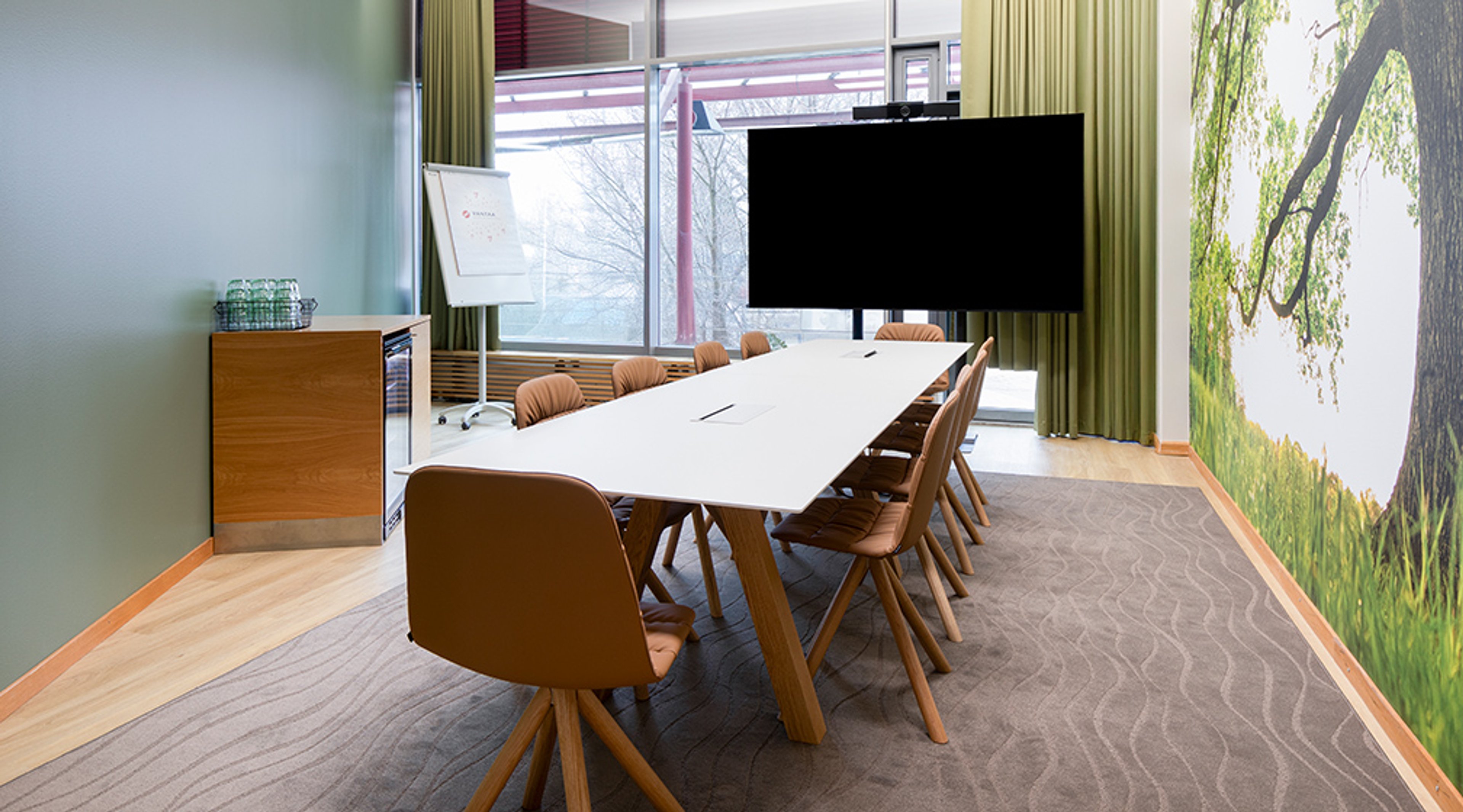 Moderni neuvotteluhuone, jossa on pitkä valkoinen pöytä, ruskeat tuolit, iso näyttö ja luontoaiheinen seinäkuva. Lattia on kovaa puuta, valaistus on hyvä.