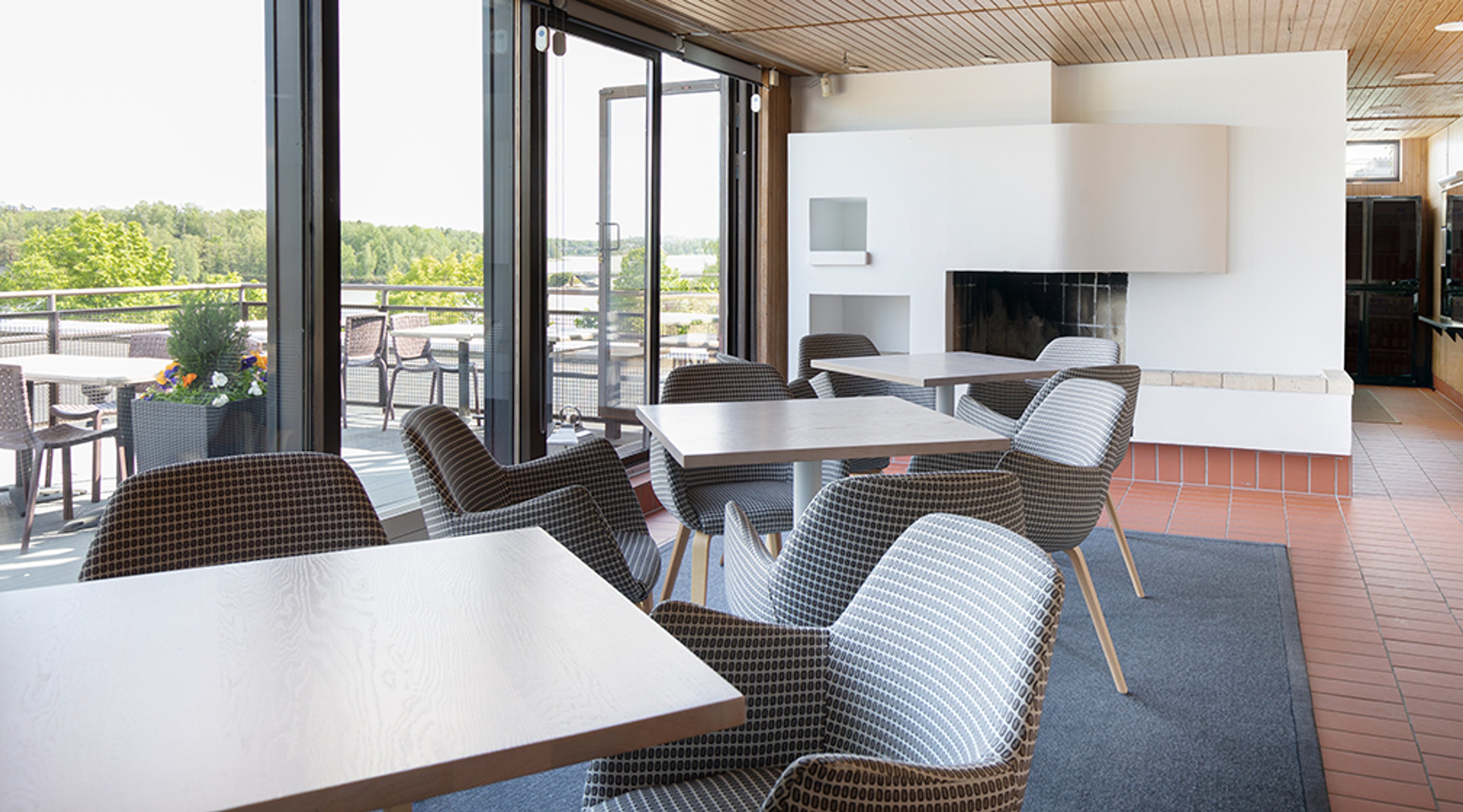 Lounge-tila, jossa moderneja pöytiä ja tuoleja, valkoinen takka ja suuret ikkunat, joista näkyy vehreä järvimaisema. Lattialla terrakottalaattoja.