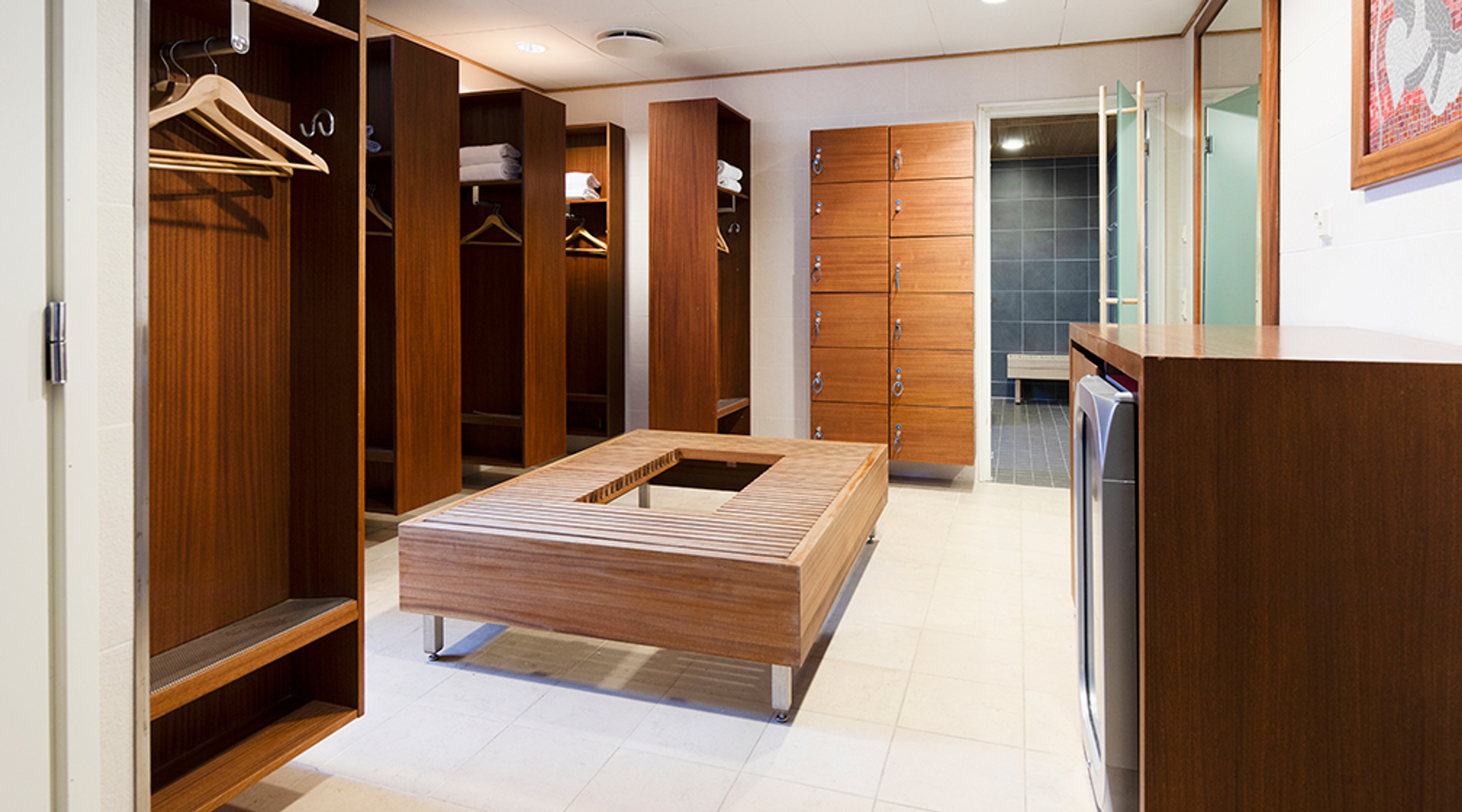 Pukuhuone puisilla lokeroilla, keskellä puinen penkki. Vasemmalla vaatteiden säilytystilaa, oikealla lukittavat kaapit. Taustalla näkyy sauna. Lattia on vaalean kivilaatoitettu.