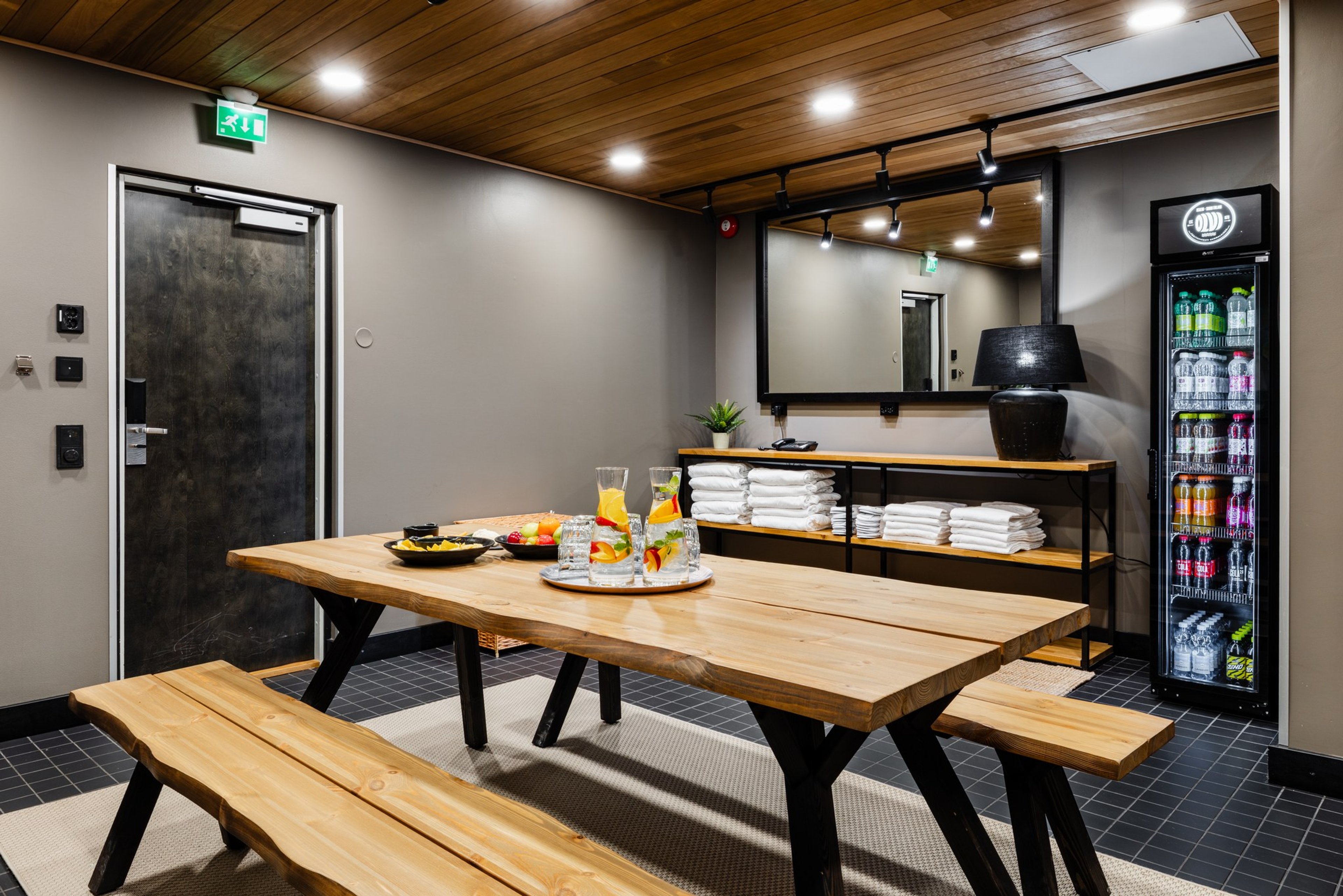 Modern sauna lounge with wooden table and benches, shelf with white towels, and beverage cooler. Floor is dark tile.