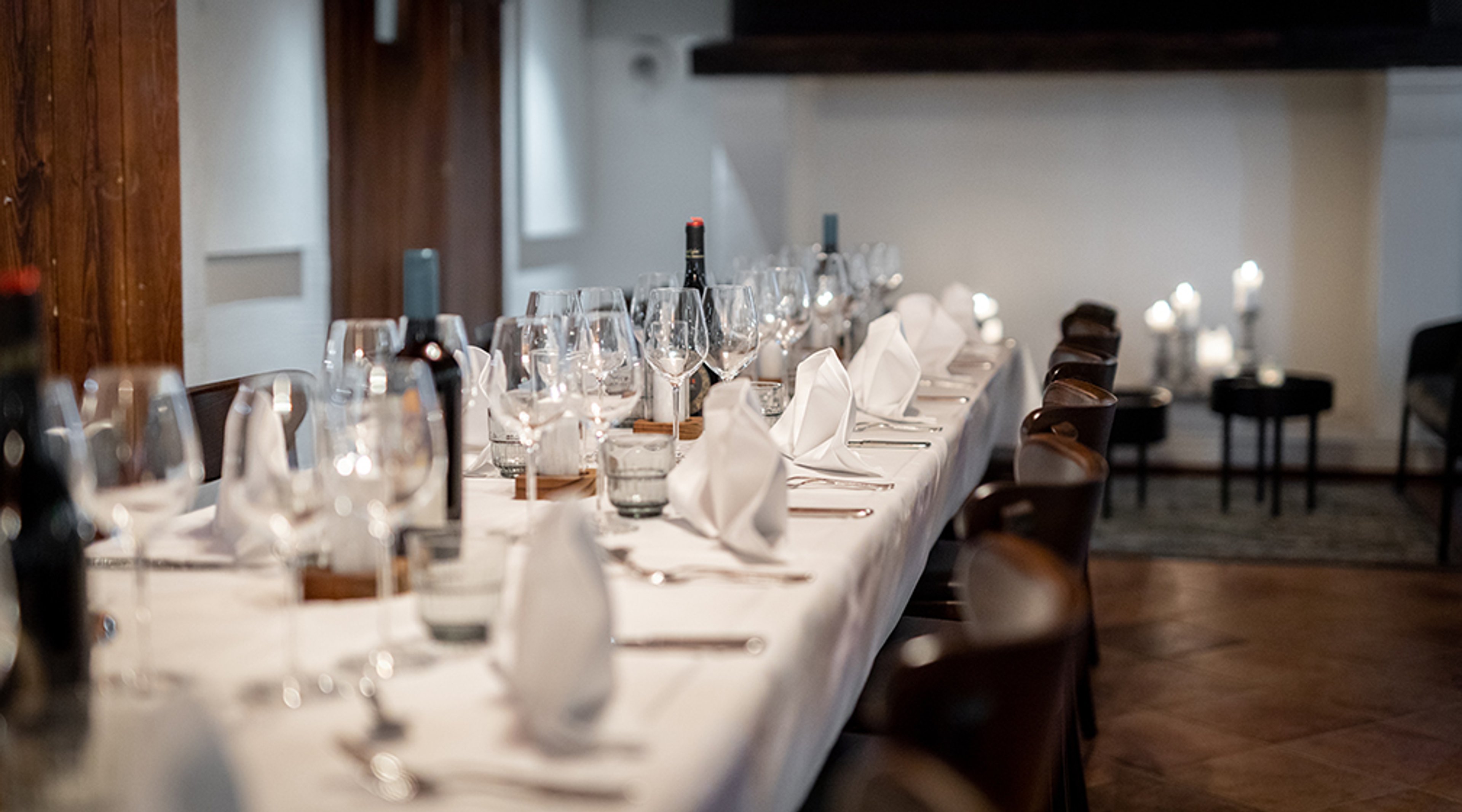 Pitkä ruokapöytä valkoisen pöytäliinan kanssa, kauniisti kattauksessa viinilaseja, taitettuja valkoisia lautasliinoja ja viinipulloja.