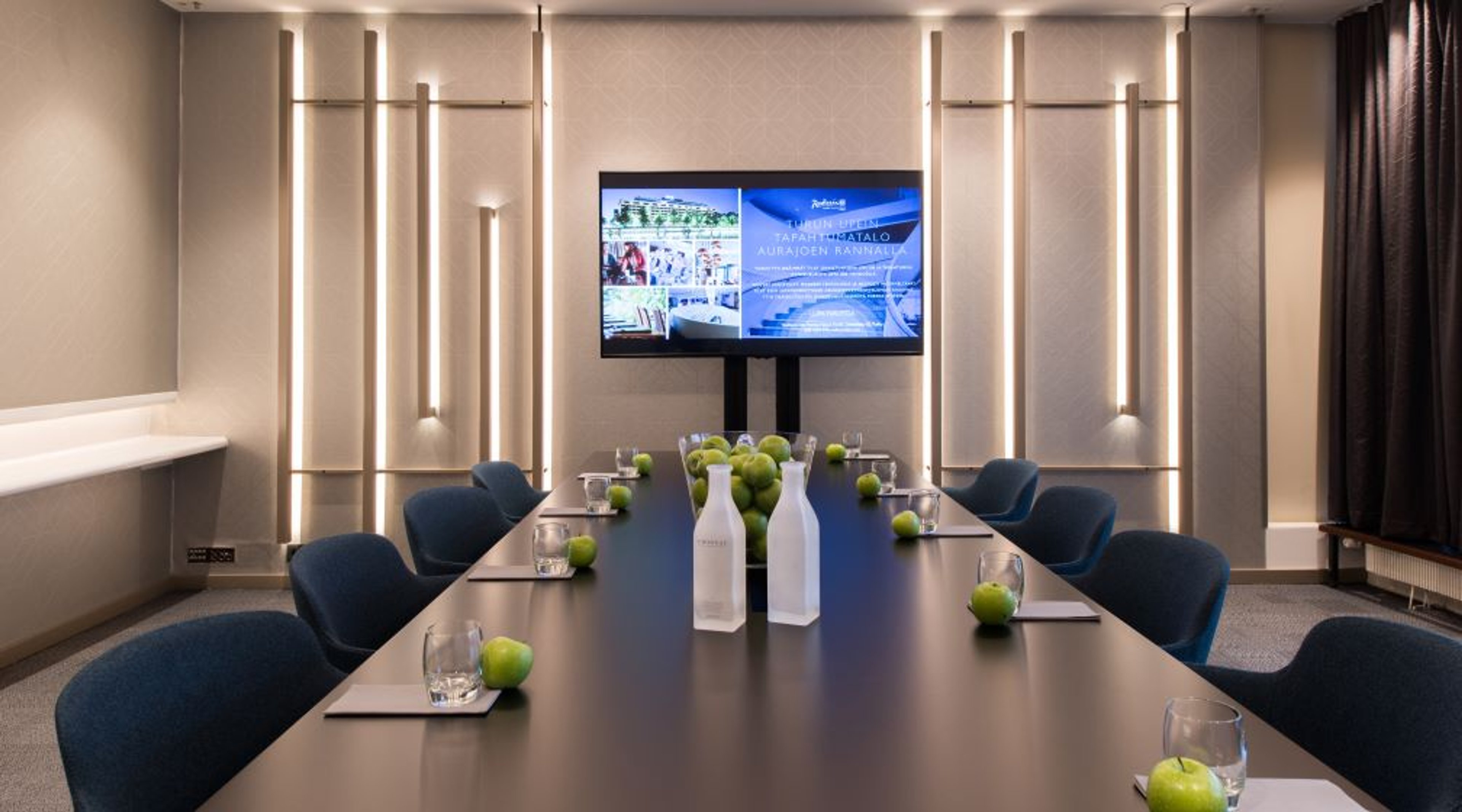 Modern conference room with dark table, blue chairs, green apples and water bottles. Wall-mounted TV screen and stylish lighting fixtures.