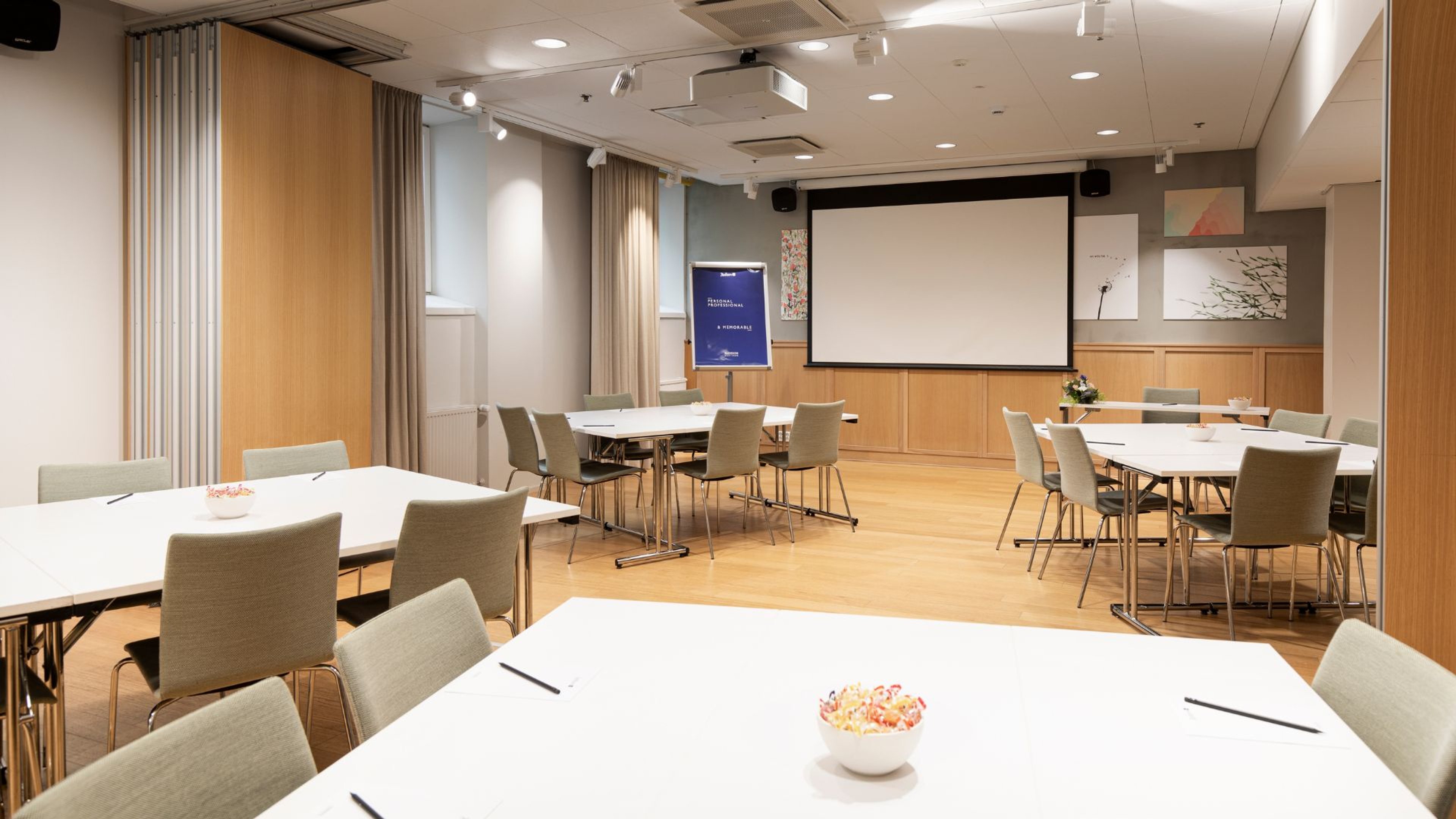 Moderni kokoustila valkoisilla pöydillä, vaaleilla tuoleilla, puisella lattialla ja projektorikankaalla. Hyvä valaistus ja esteetön lattia.
