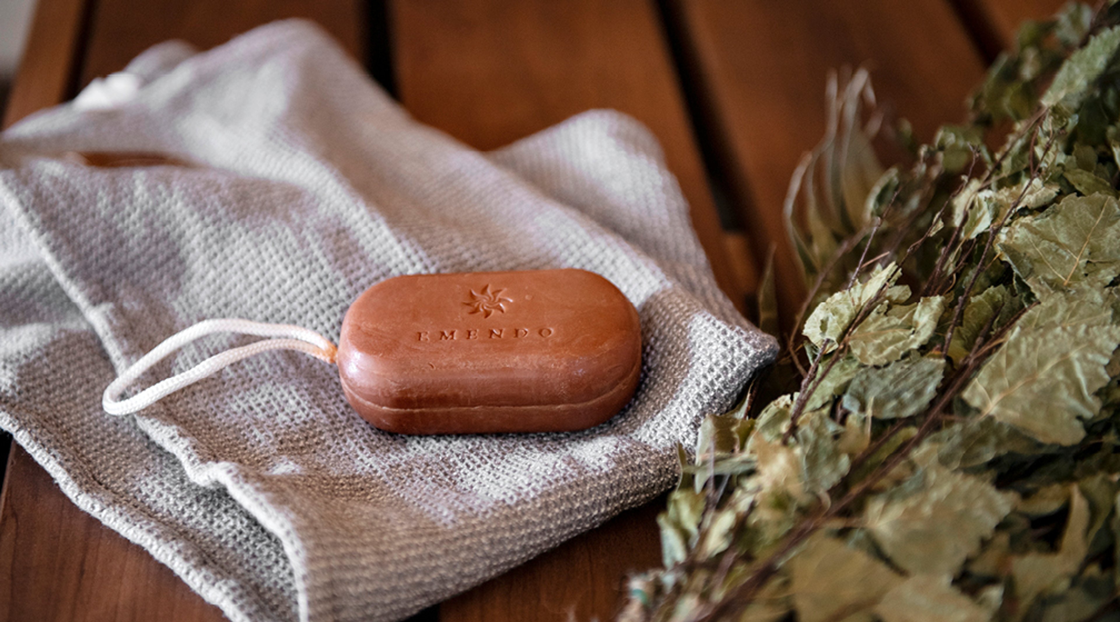 Ruskea kappale saunatuotetta vaalean pyyhkeen päällä puisella alustalla, vieressä kuivattuja saunavihtoja.