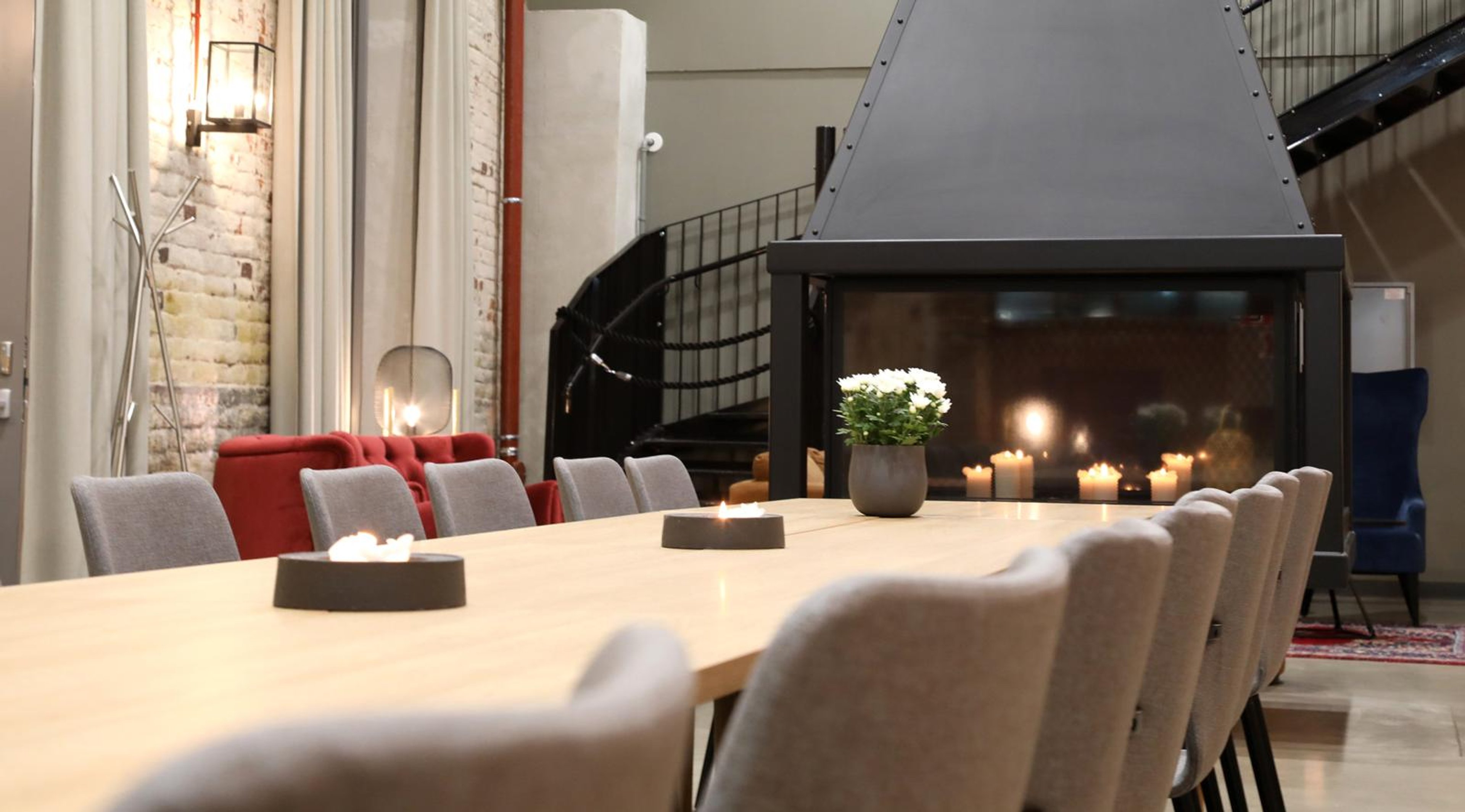 Tyylikäs kokoushuone pitkällä puupöydällä, harmailla tuoleilla ja modernilla takkalla. Punaiset sohvat ja tiiliseinä luovat lämpimän tunnelman.