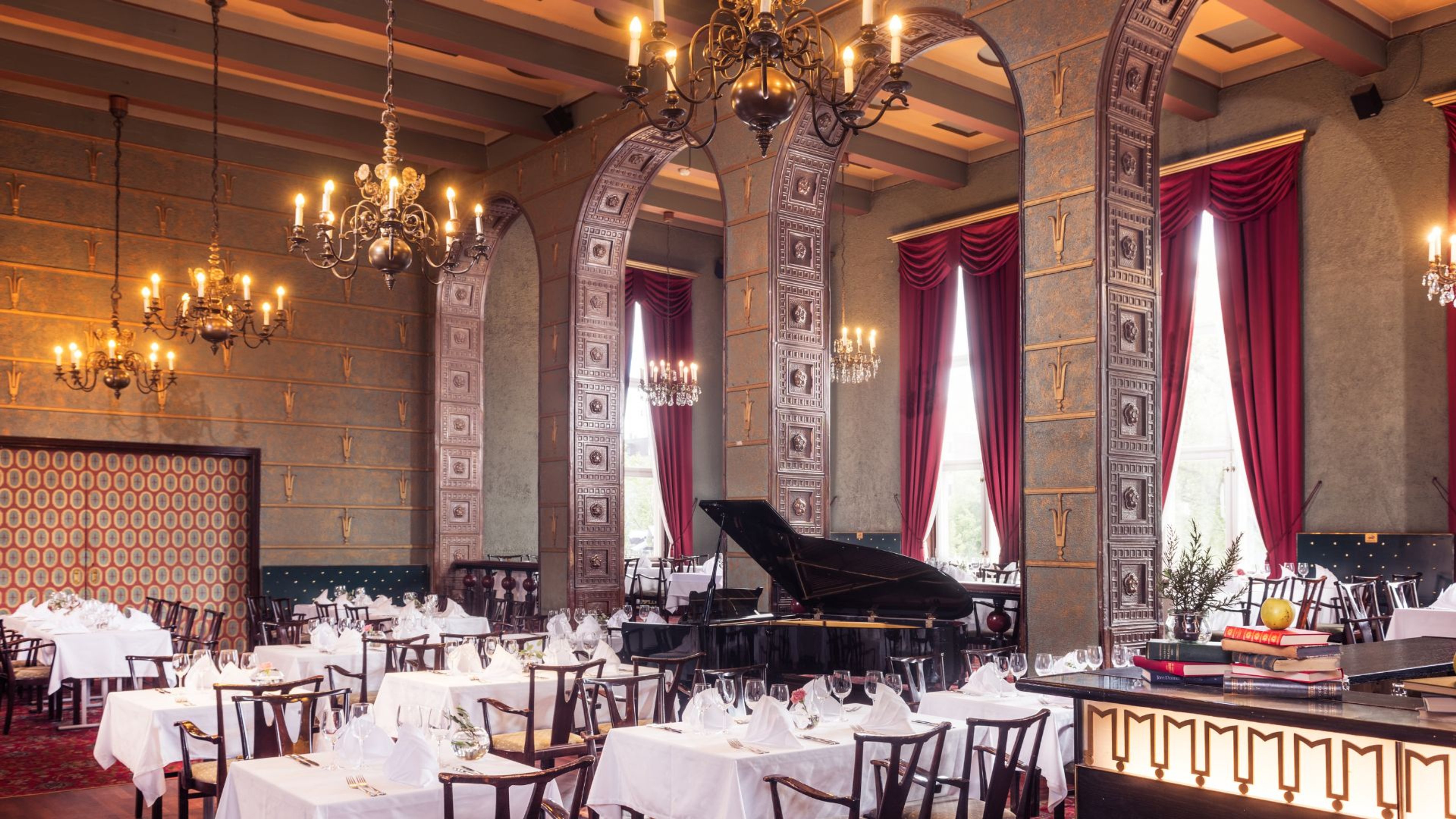 Elegantti ravintolasali koristeellisilla kaarilla, kattokruunuilla ja punaisilla verhoilla. Keskellä on flyygeli, ympärillä valkoliinaiset pöydät.
