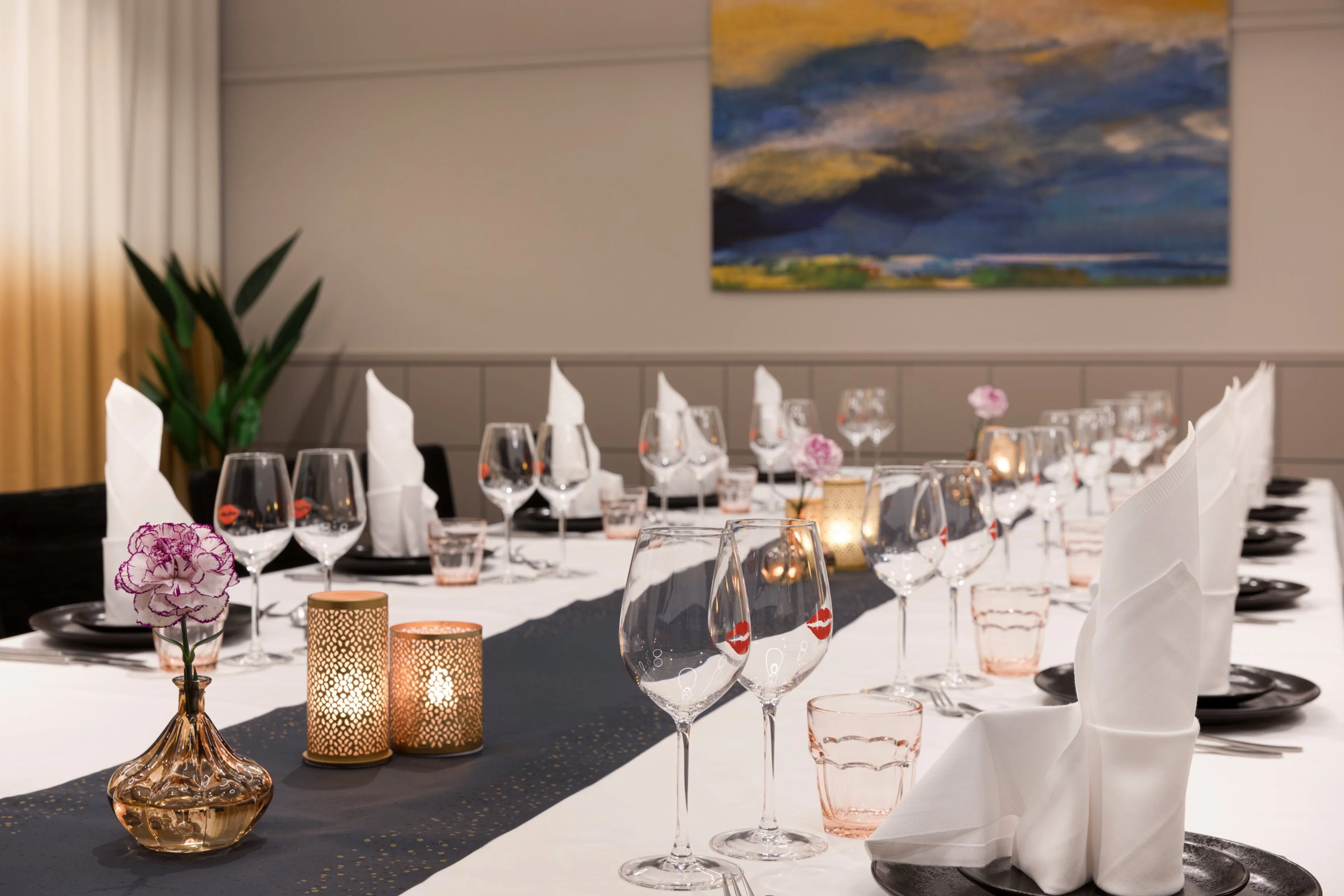 Elegantisti katettu pitkä pöytä ravintolassa. Valkoisella liinalla, mustilla lautasilla ja viinilaseilla. Pöydällä on kynttilöitä ja kukkakoristeita. Taustalla sinisävyinen taideteos.