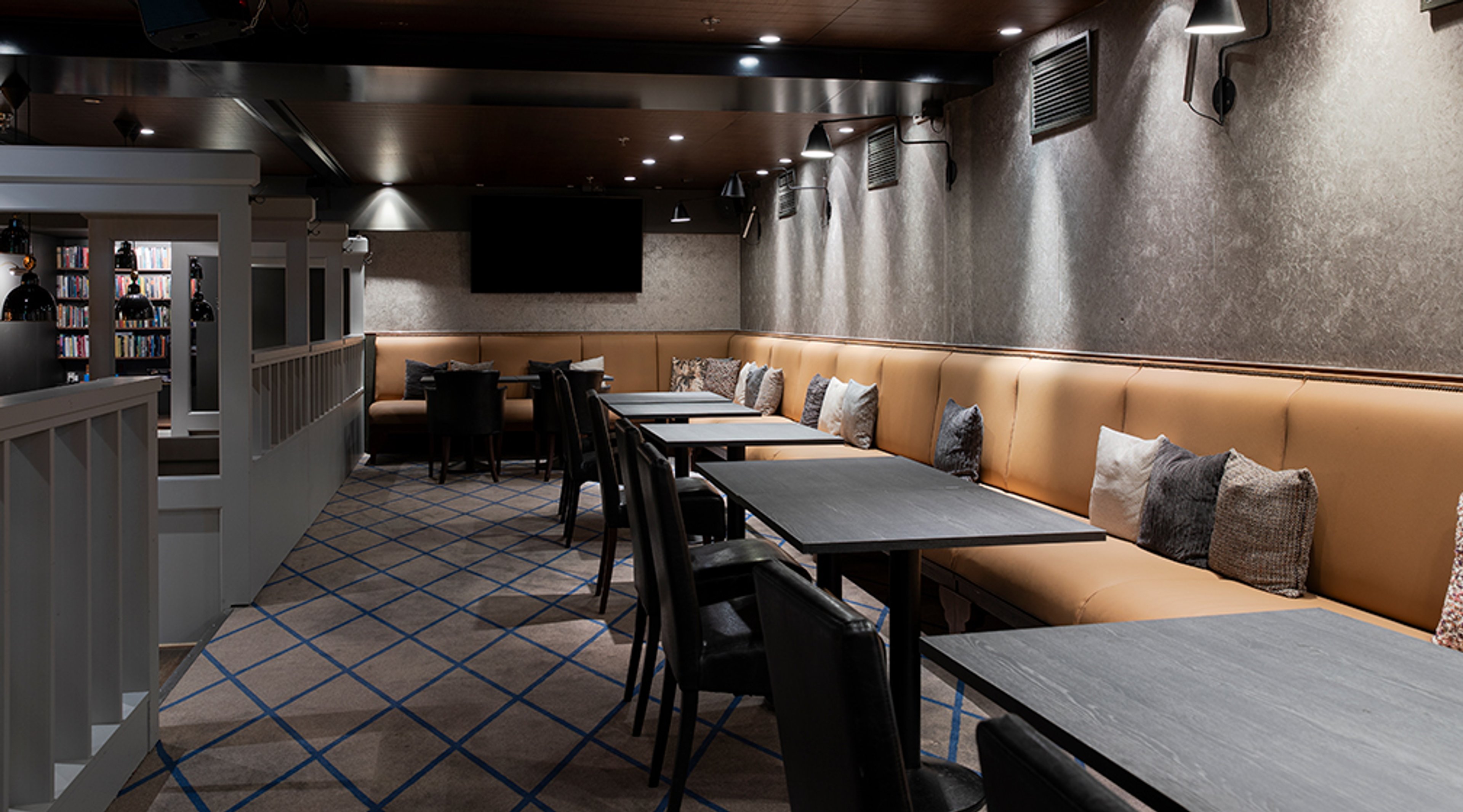 Ravintolan lounge-tila, jossa on oranssi sohva, mustia pöytiä ja tuoleja, sekä kuvioitu lattia. Seinällä TV ja kattovalaistus.