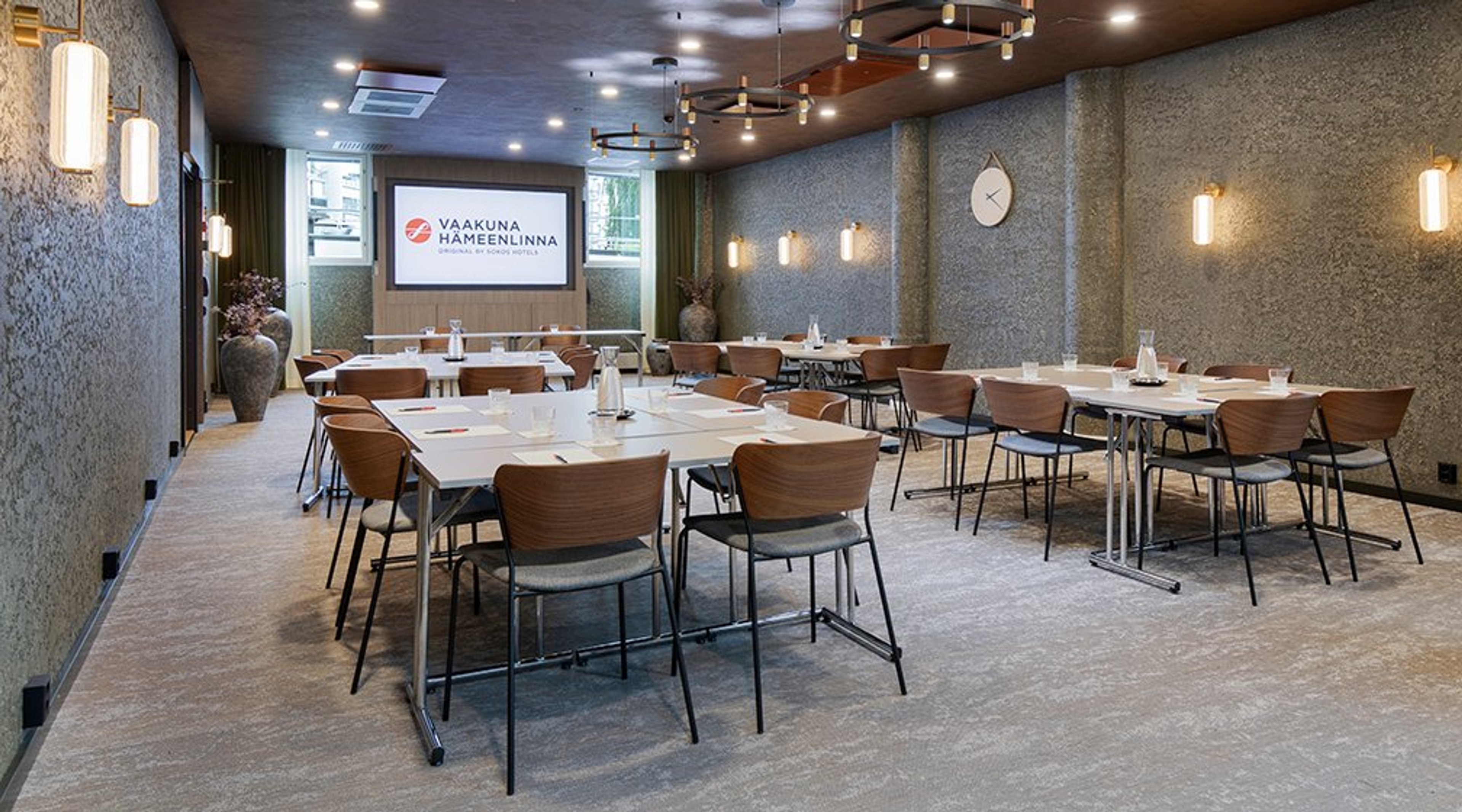 Moderni kokoustila, jossa pöytiä ja puuselkäisiä tuoleja. Taustalla näkyy valkokangas. Tila on valaisttu seinälampuilla ja kattovaloilla. Lattia on tasainen.