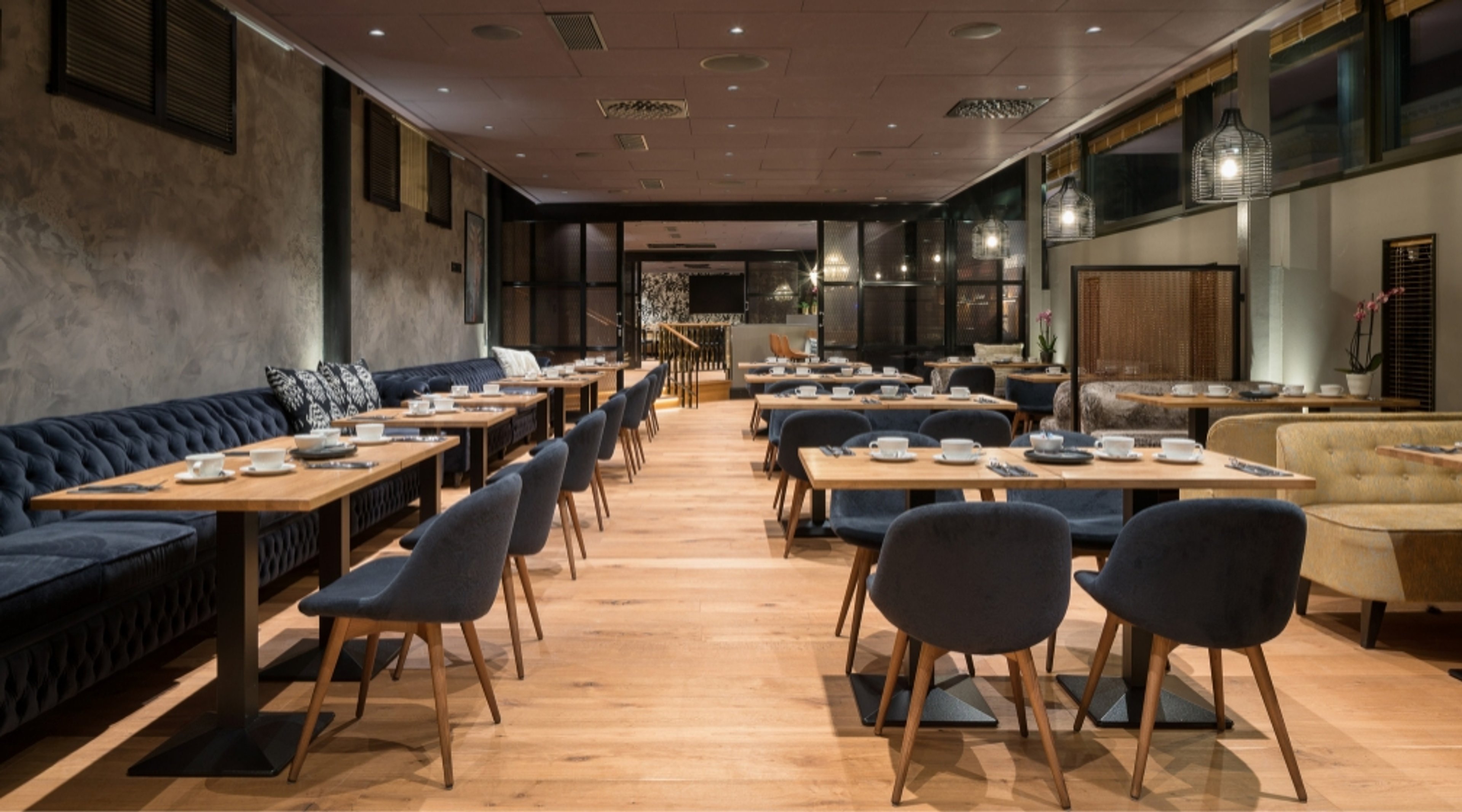 Tyylikkäästi sisustettu ravintola puulattialla, sinisillä tuoleilla ja sohvilla sekä puupöydillä. Tilassa häkkimäiset valaisimet ja tasainen valaistus.