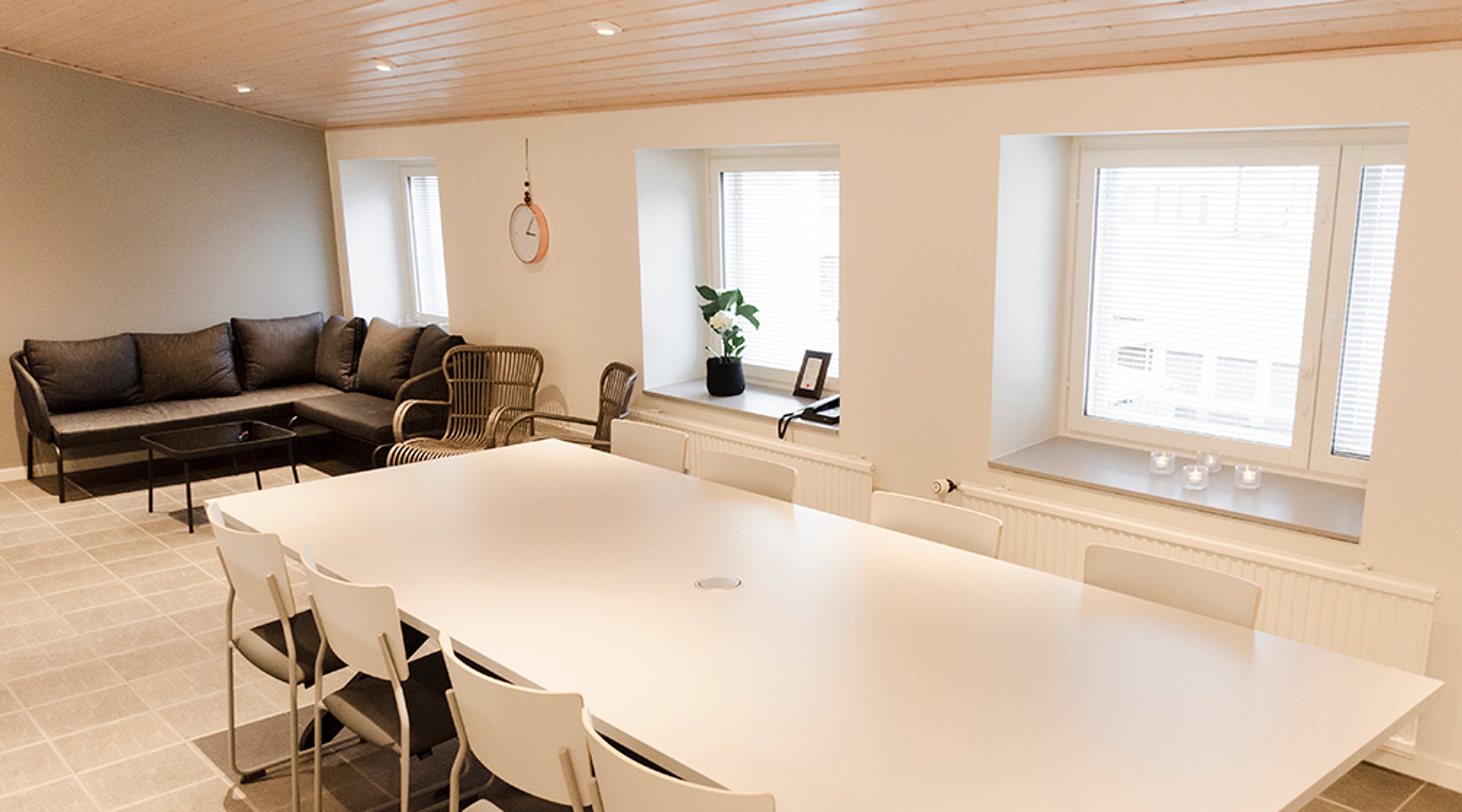 Valoisa neuvotteluhuone, jossa on valkoinen pöytä tuoleineen, tumma sohvaryhmä ja rottinkituolit. Kolme suurta ikkunaa ja puupaneloitu katto.