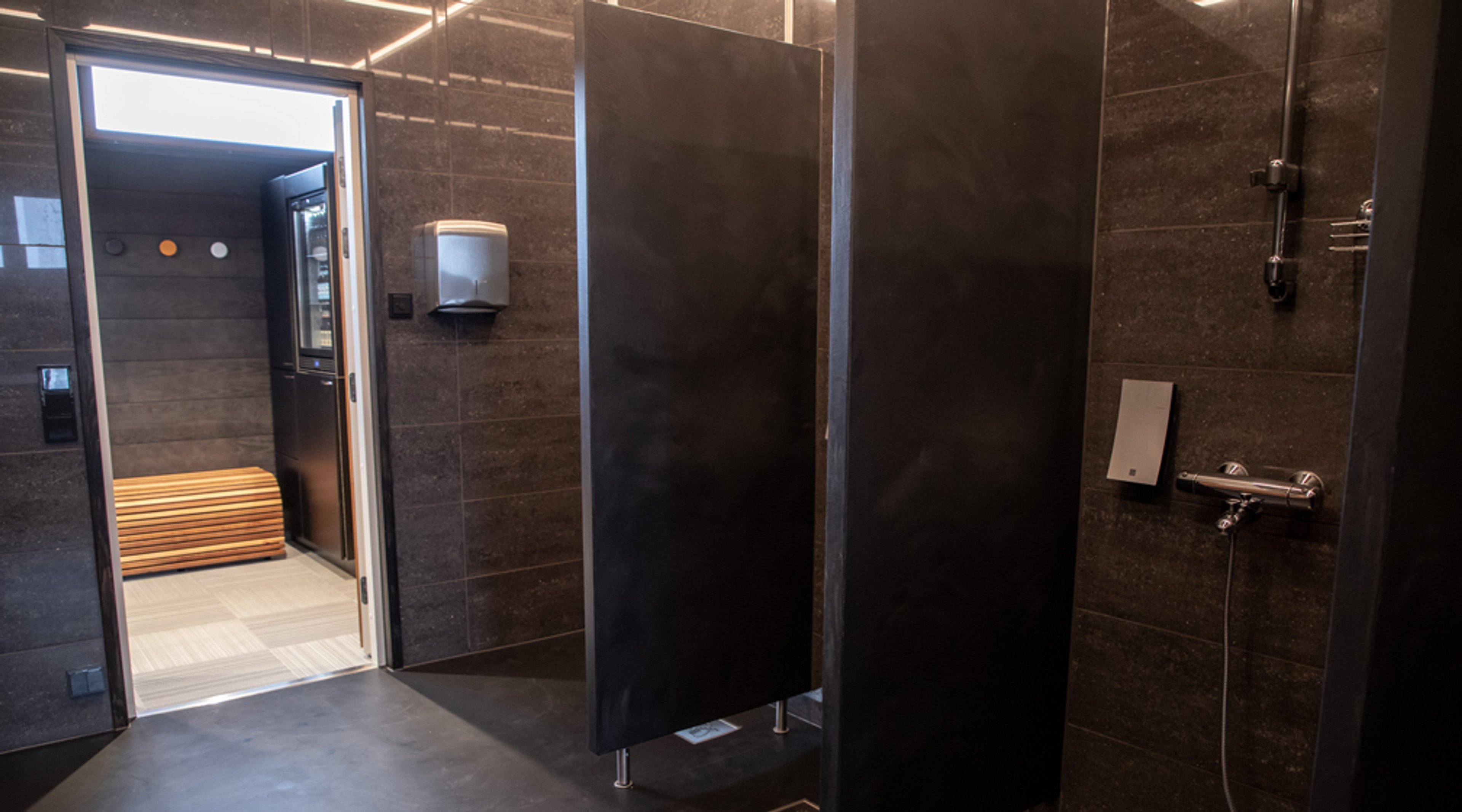 Saunan pukuhuone tummilla laatoilla, suihkut ja erilliset pukukaapit. Avoin ovi saunatilaan, jossa näkyy puinen laude.