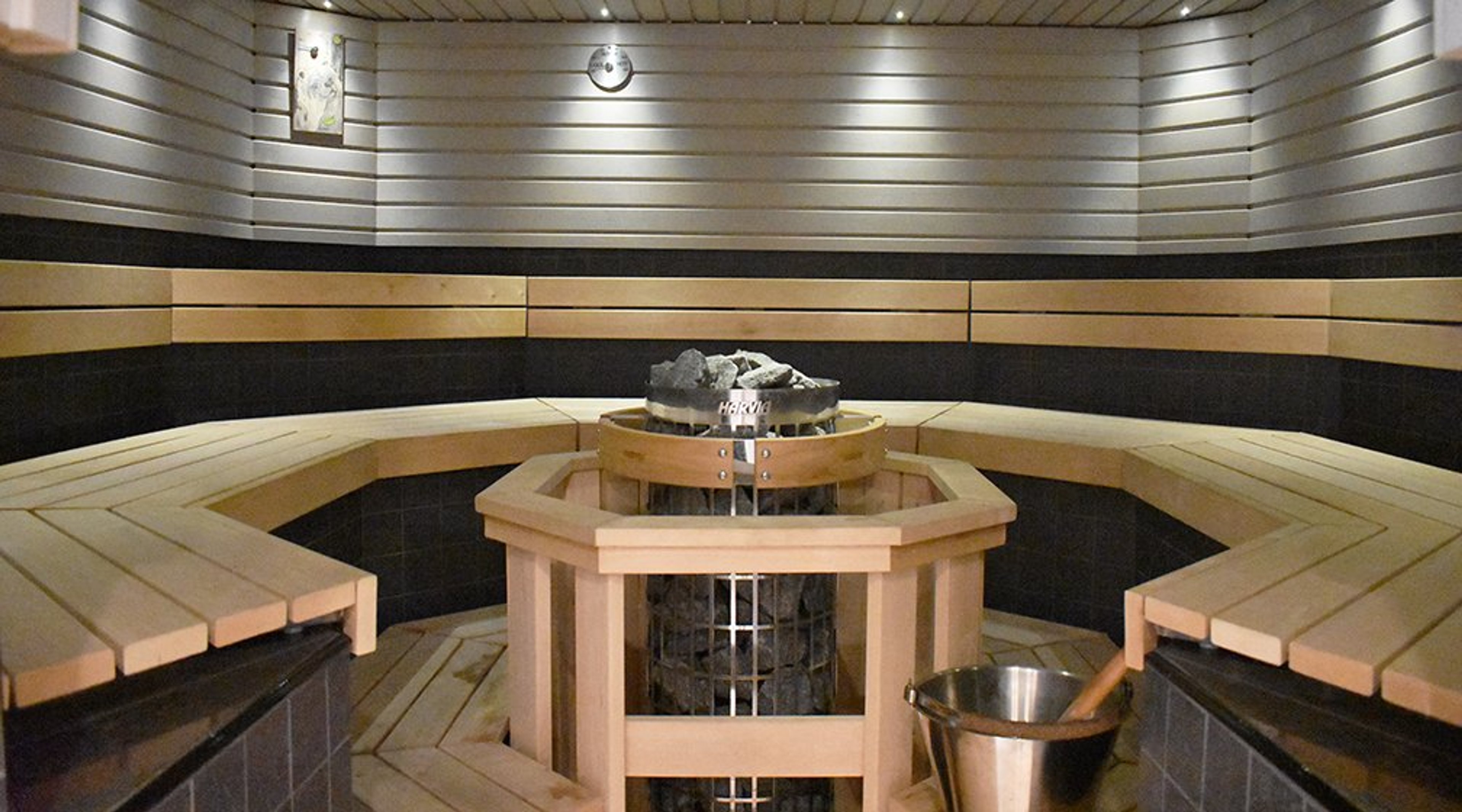 Moderni sauna puisilla lauteilla ja kuusikulmaisella kiukaalla keskellä. Vaaleat puupaneelit koristavat seinäpintaa.